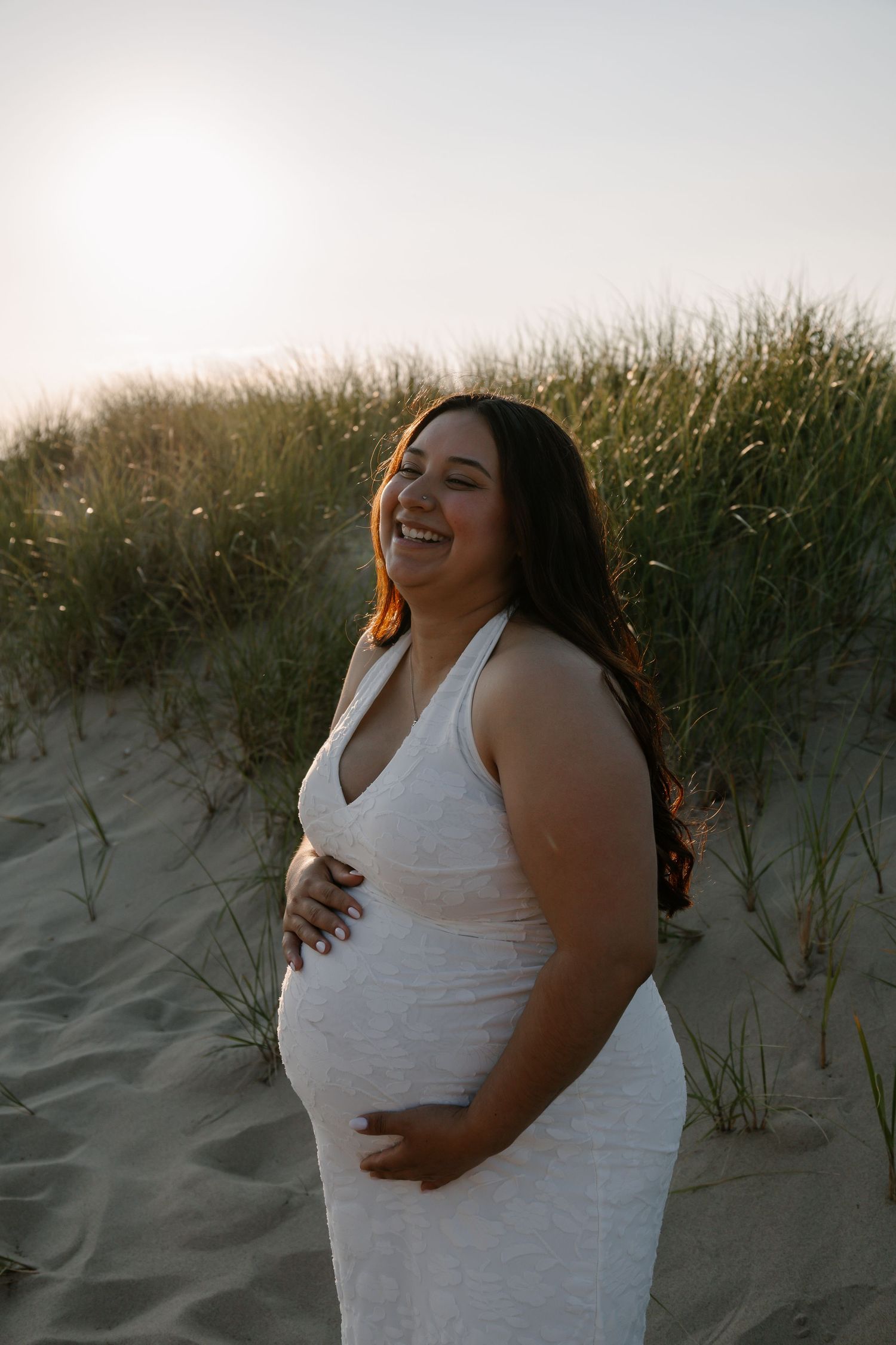 Smiling pregnant woman in white dress holding belly on sandy beach dunes at golden hour sunset.