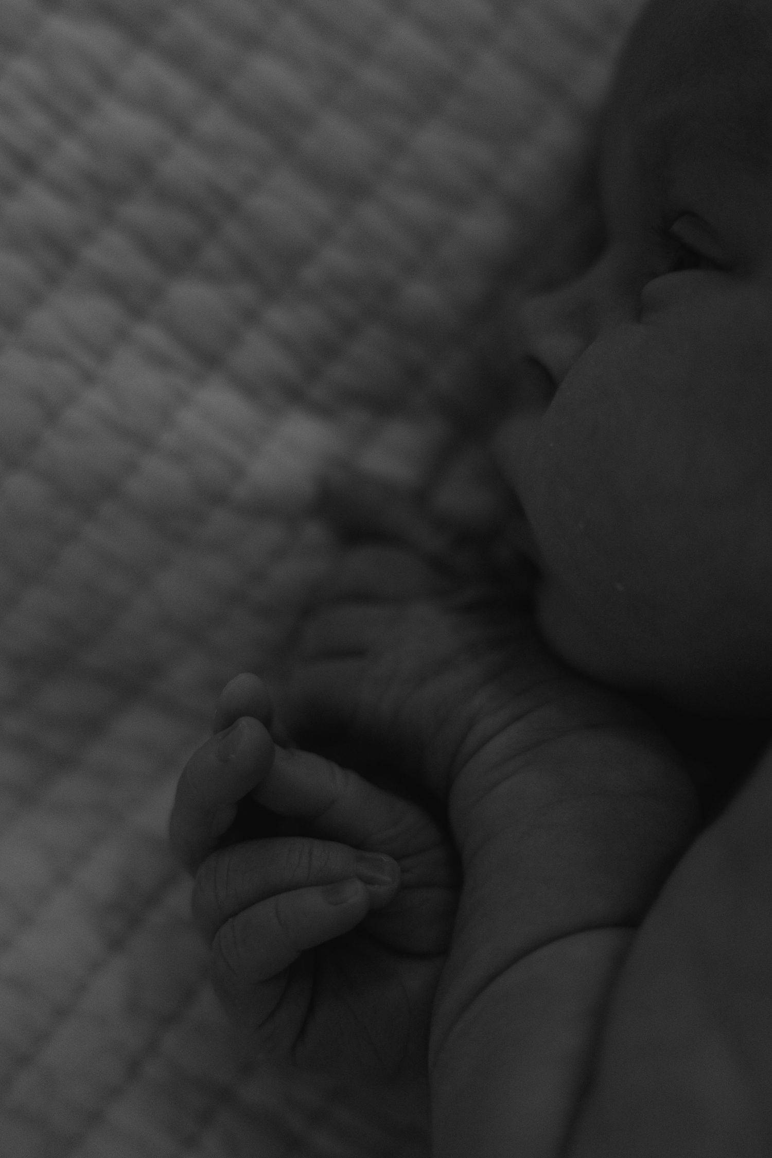 Black and white close-up photo of a sleeping newborn baby with tiny hands curled near face on quilted blanket.