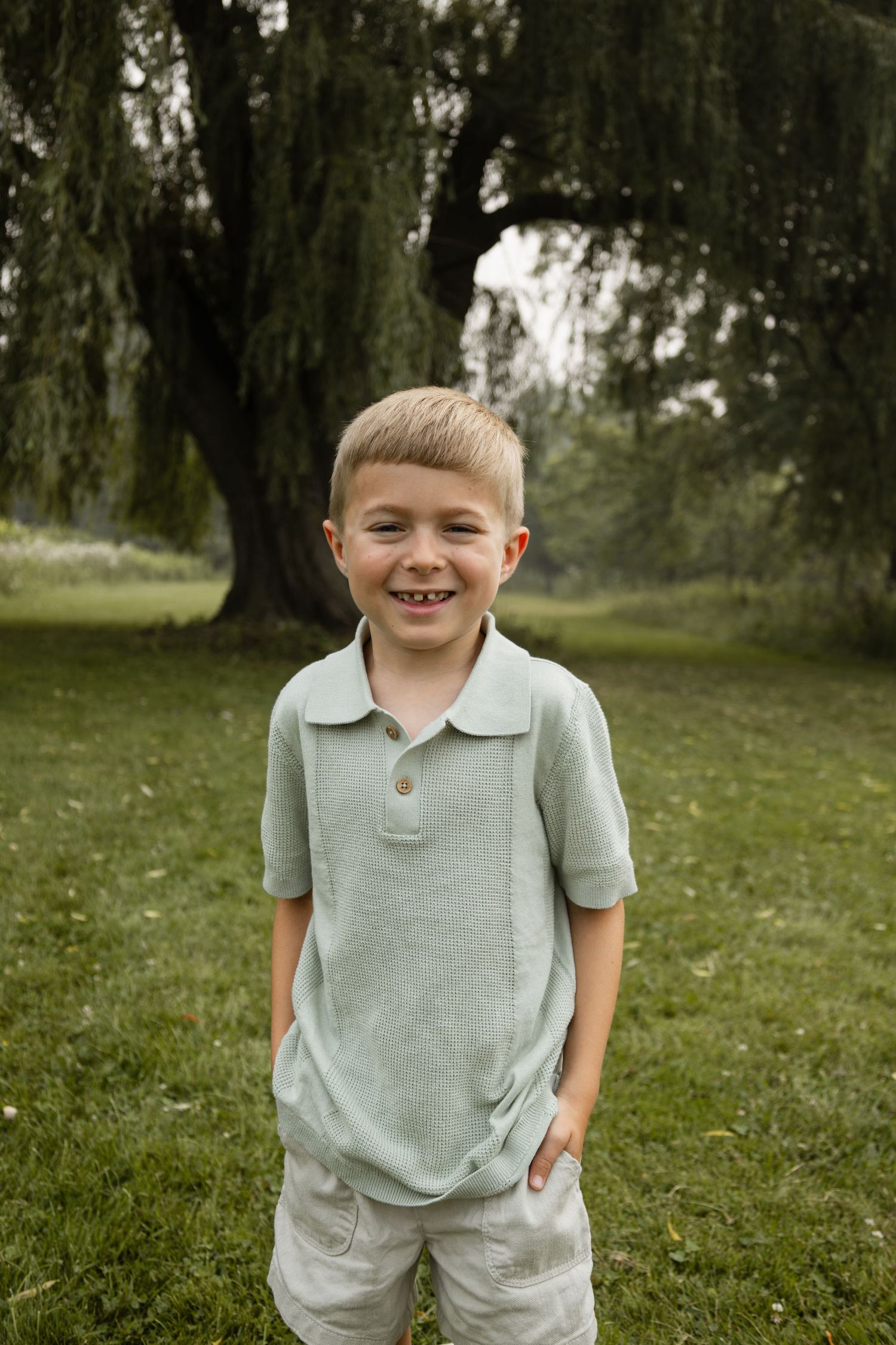 Happy blonde boy smiling with hands in pockets standing in green park by willow tree.