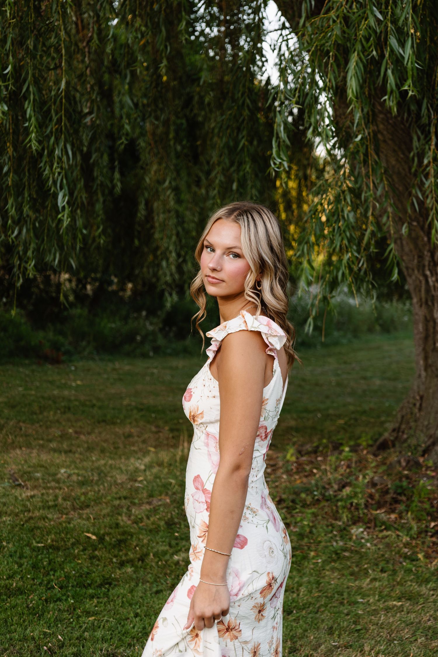 Young woman in floral dress posing outdoors under weeping willow tree on lush green lawn.
