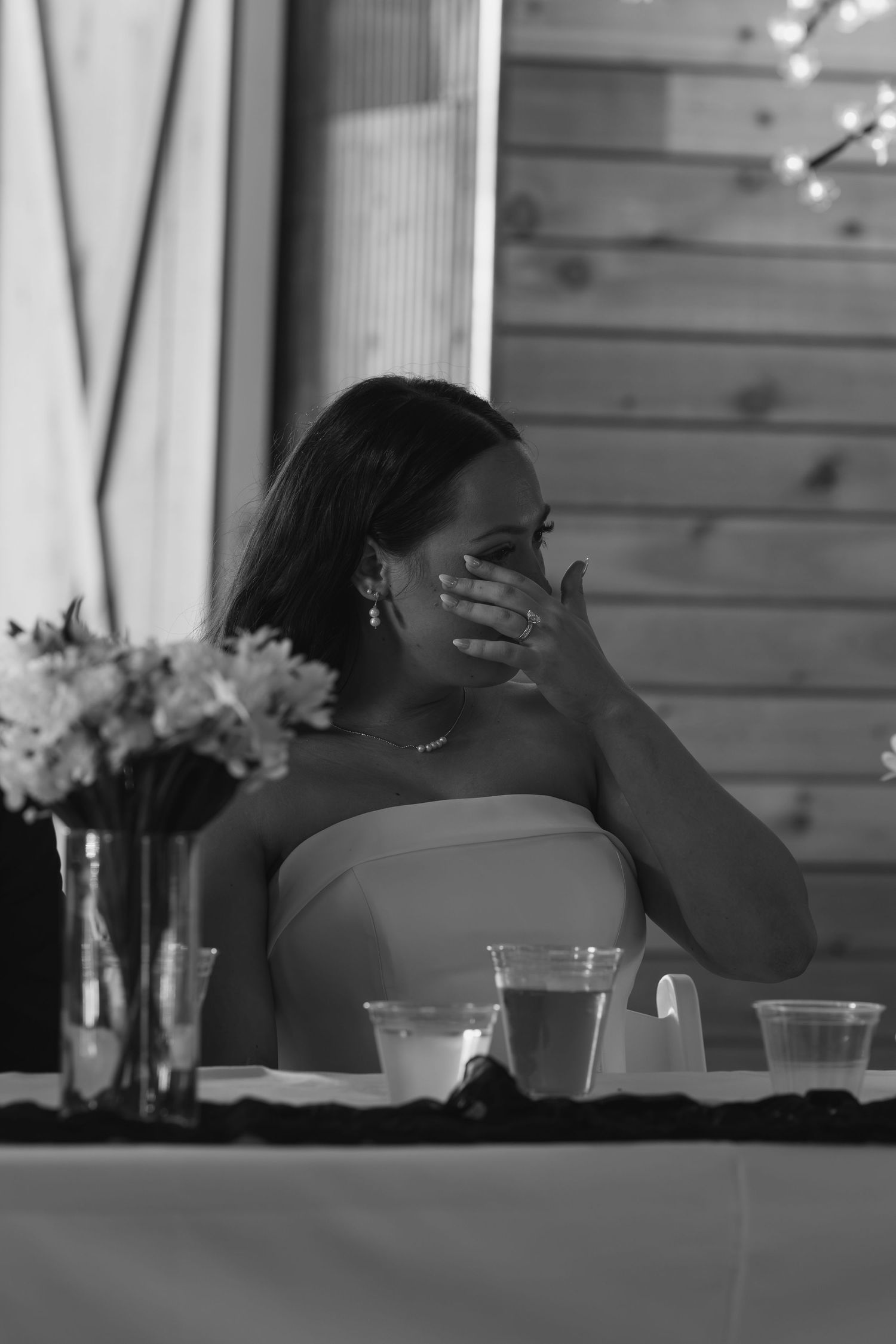 Emotional woman in strapless dress covers face with hand at wedding reception table.