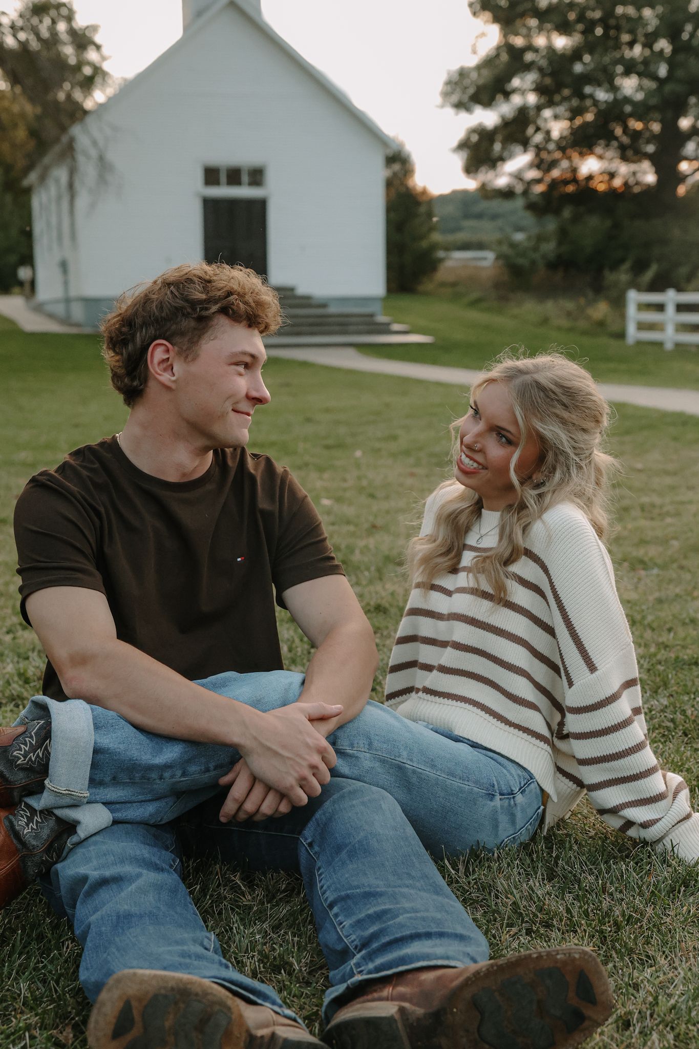 A couple sits together on green grass, smiling at each other with a white country church in the background.