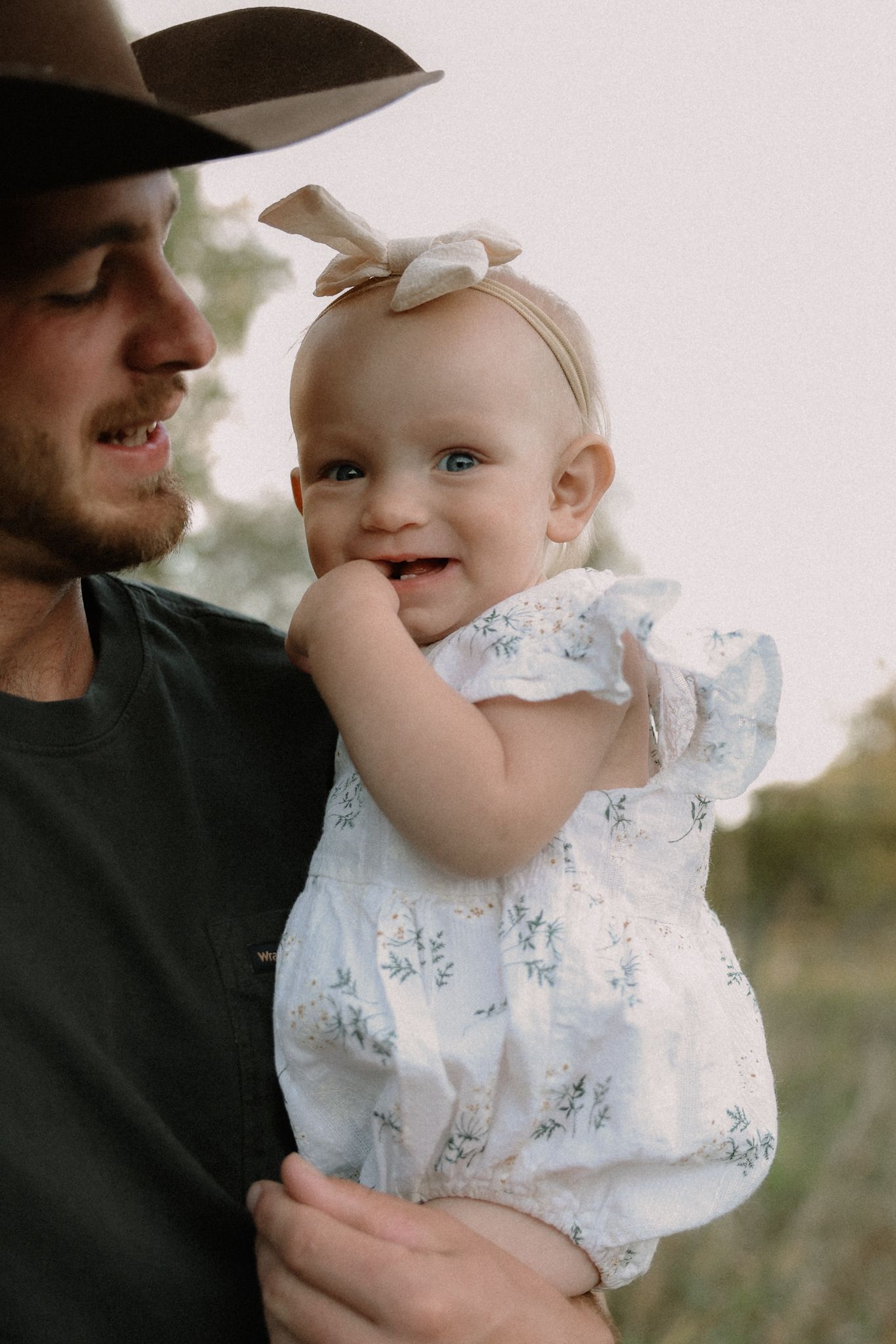 A smiling baby girl in a floral romper and bow headband being held by a man in a cowboy hat outdoors.