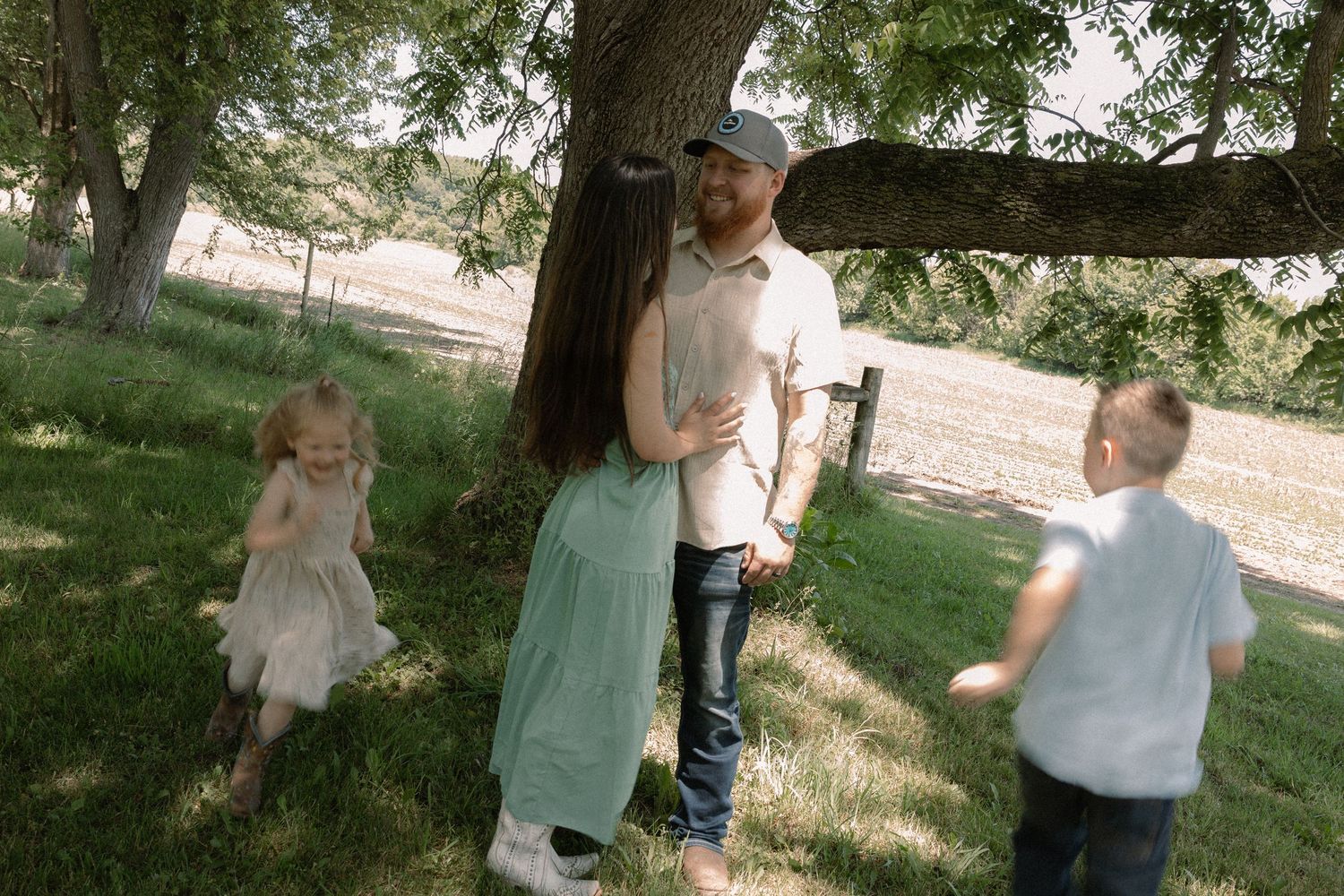 Couple sharing a romantic moment under a tree while two young children play around them outdoors.
