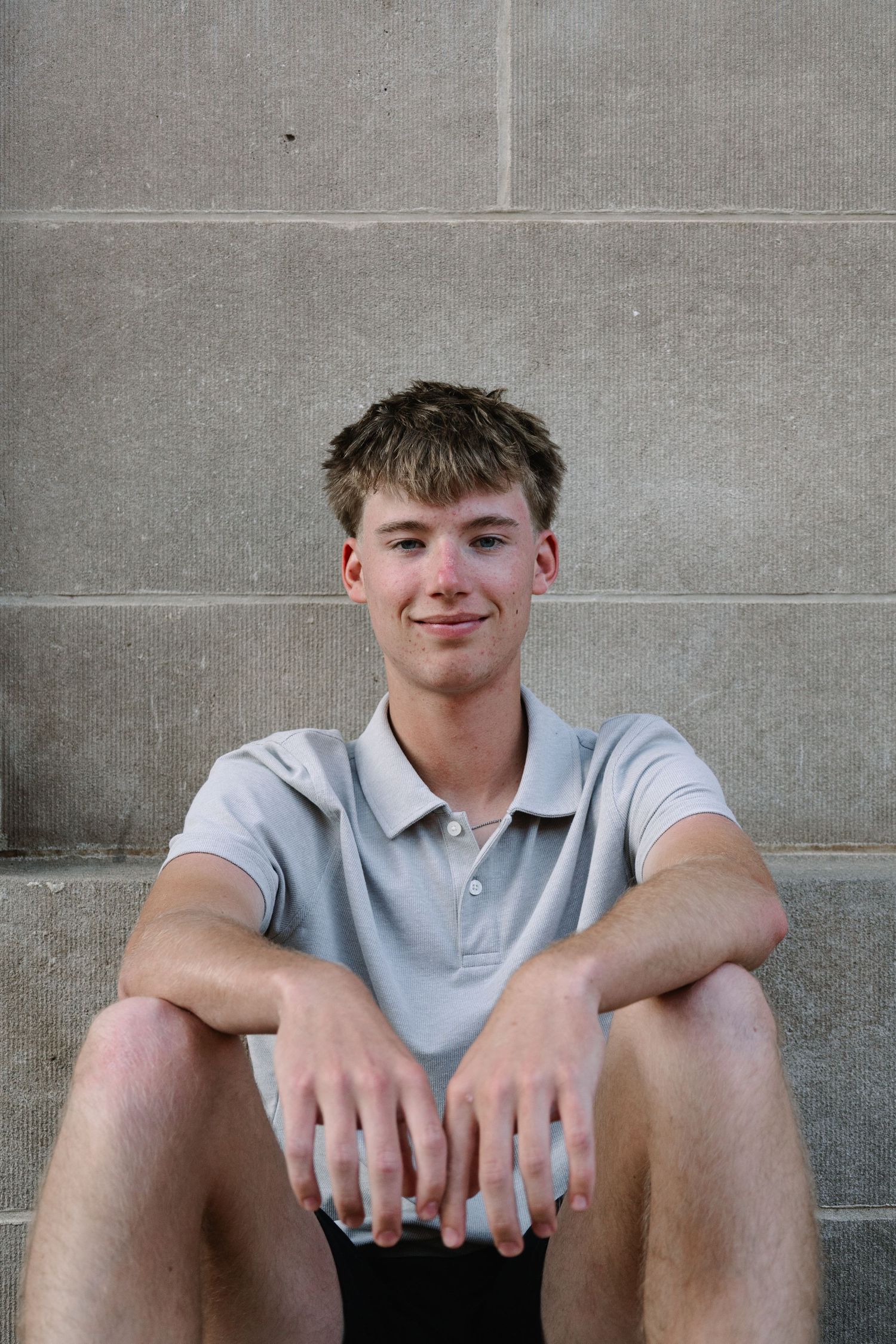 Young man in gray polo shirt sitting against stone wall, smiling confidently at camera.