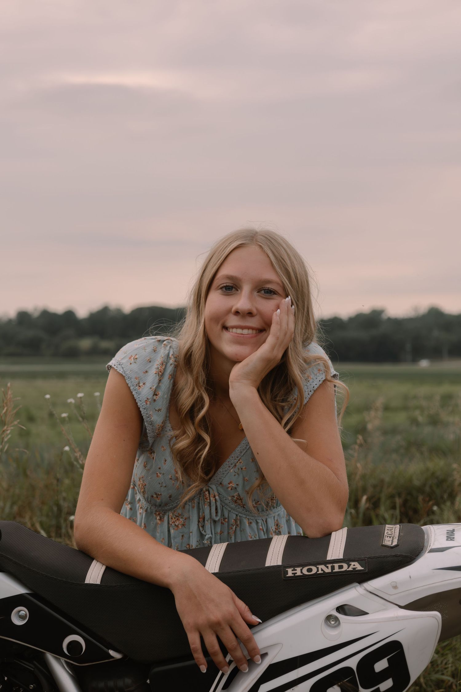 Smiling blonde woman in floral dress leaning on a Honda dirt bike in a green field at dusk.