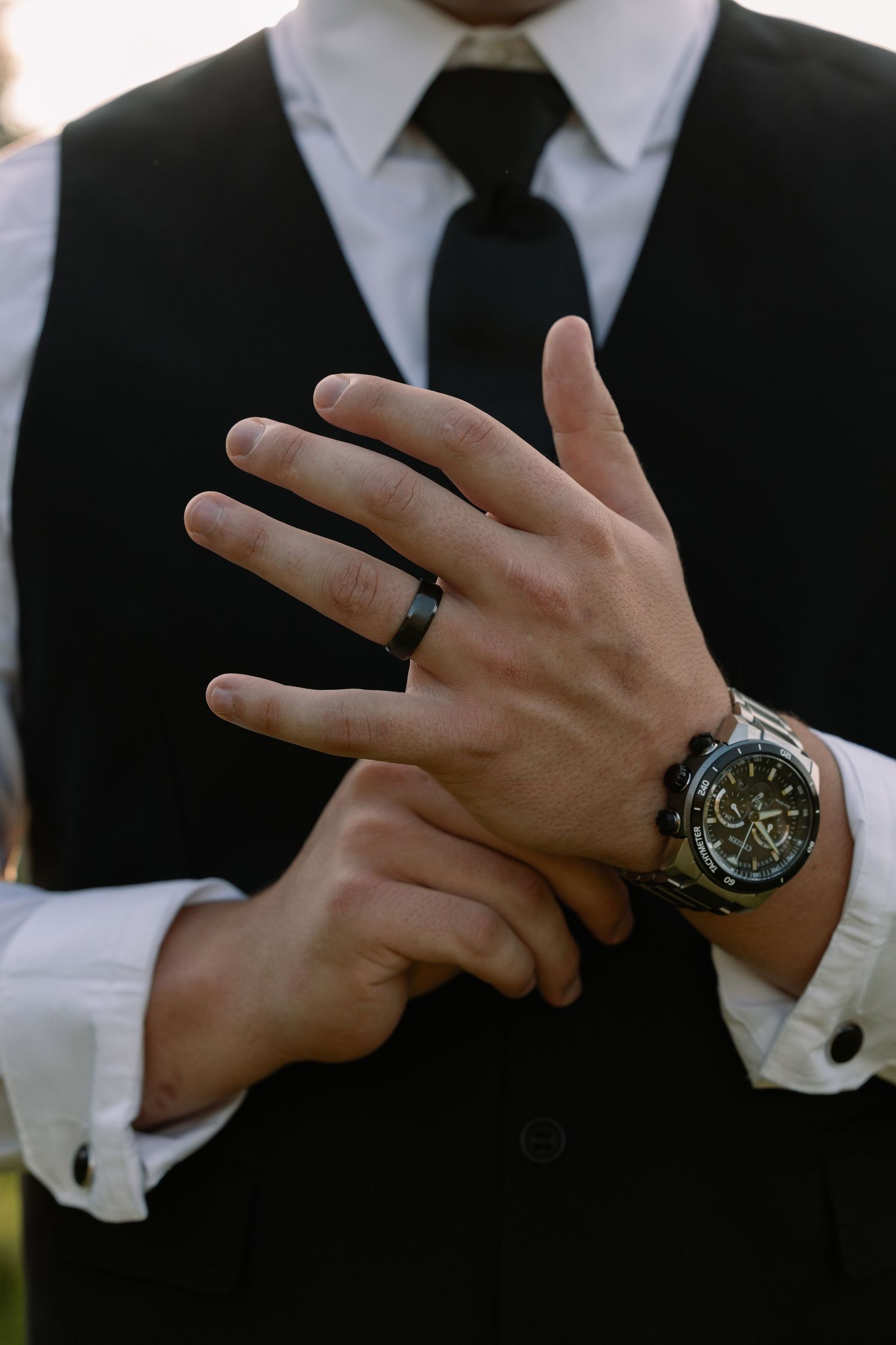Groom in black vest and tie displays wedding ring and luxury watch on his hands.