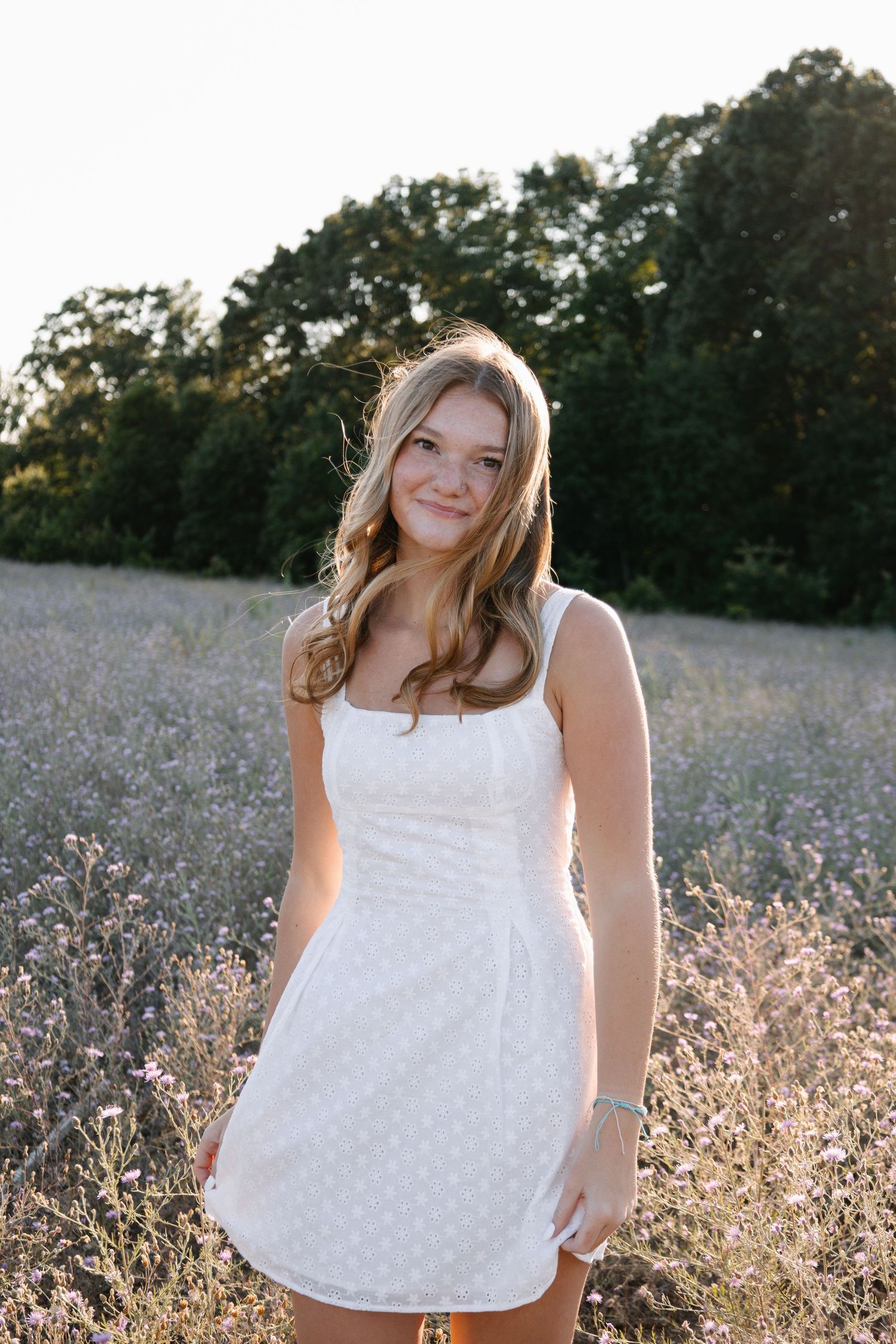Young woman in white sundress smiling in a wildflower field at golden hour with trees in background.