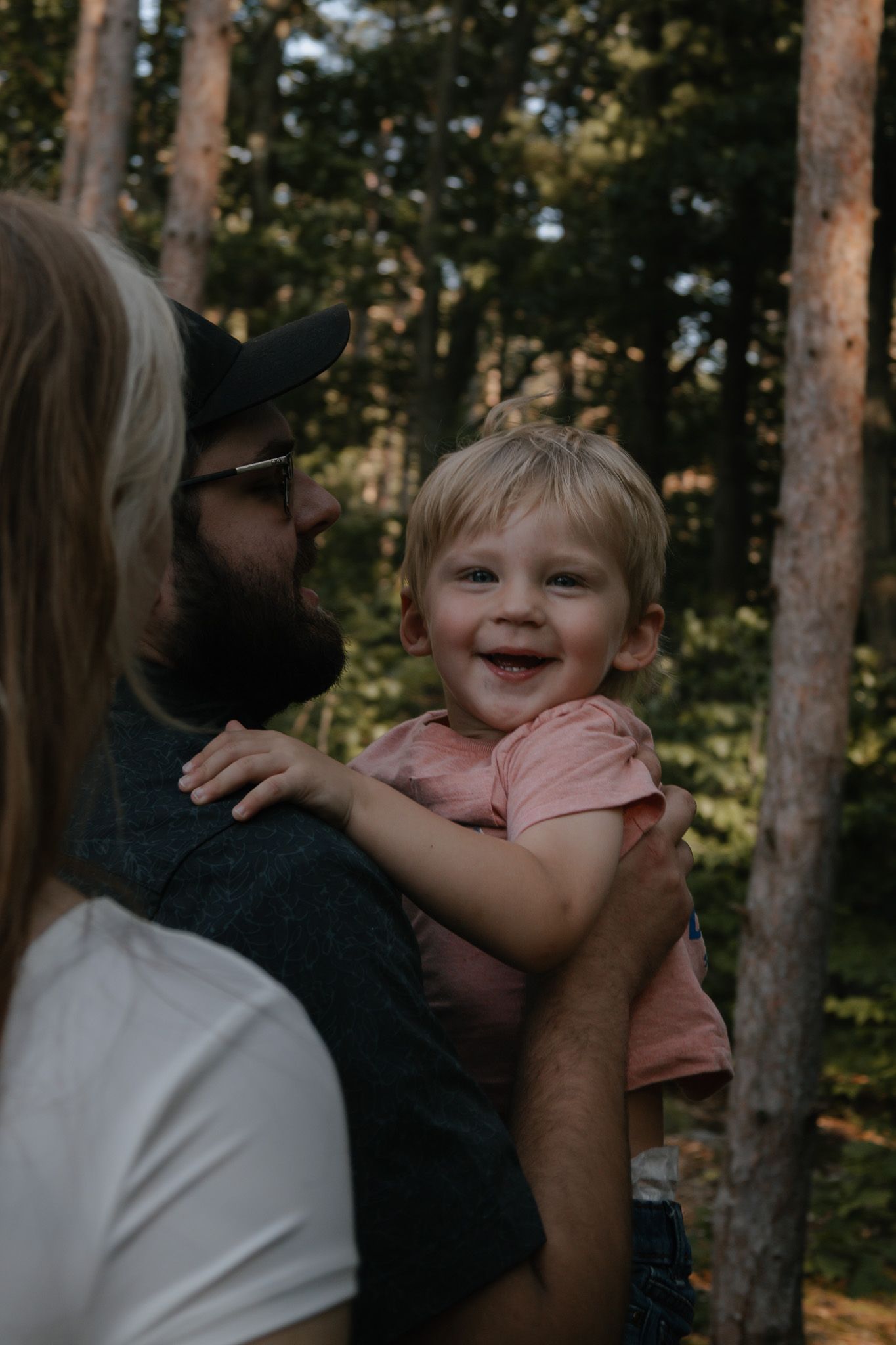A bearded man in a cap holds a smiling toddler in a pink shirt outdoors in a sunlit forest.