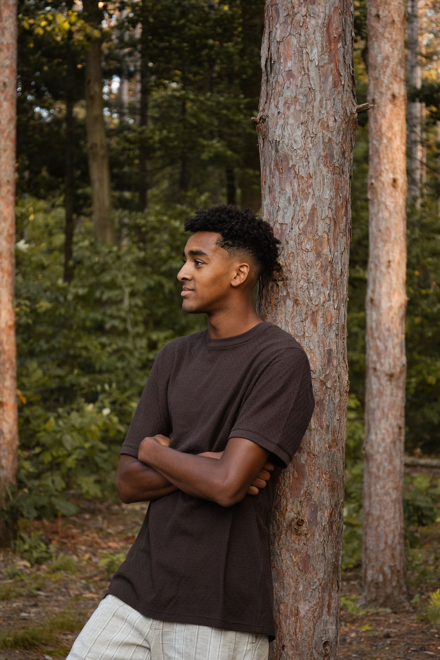 Young Black man in brown t-shirt leaning against pine tree in forest with arms crossed, looking sideways.