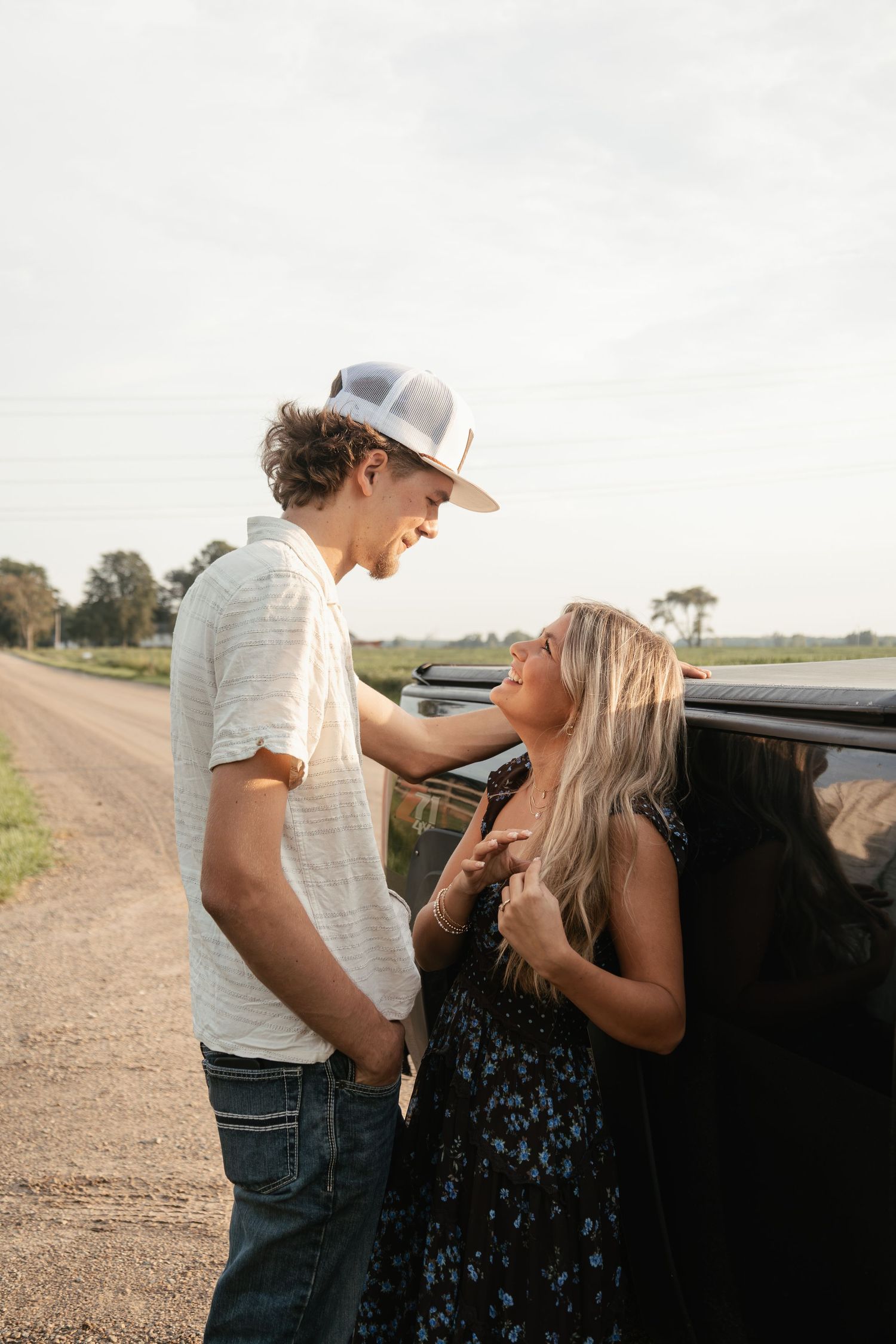 A young couple shares a romantic moment beside a black truck on a rural dirt road at golden hour.