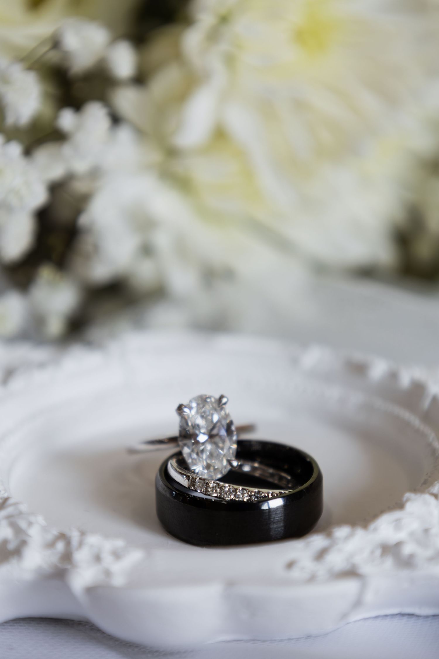 Wedding rings set on white dish with diamond engagement ring and black band, white floral bouquet in background.