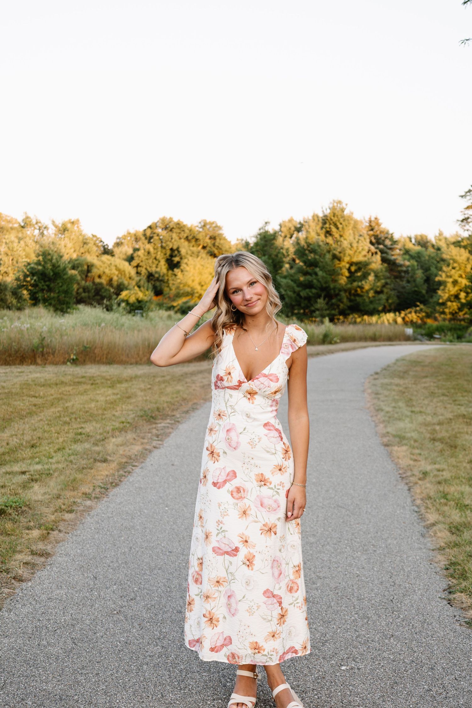 Blonde woman in floral maxi dress standing on a park path surrounded by lush green trees at sunset.