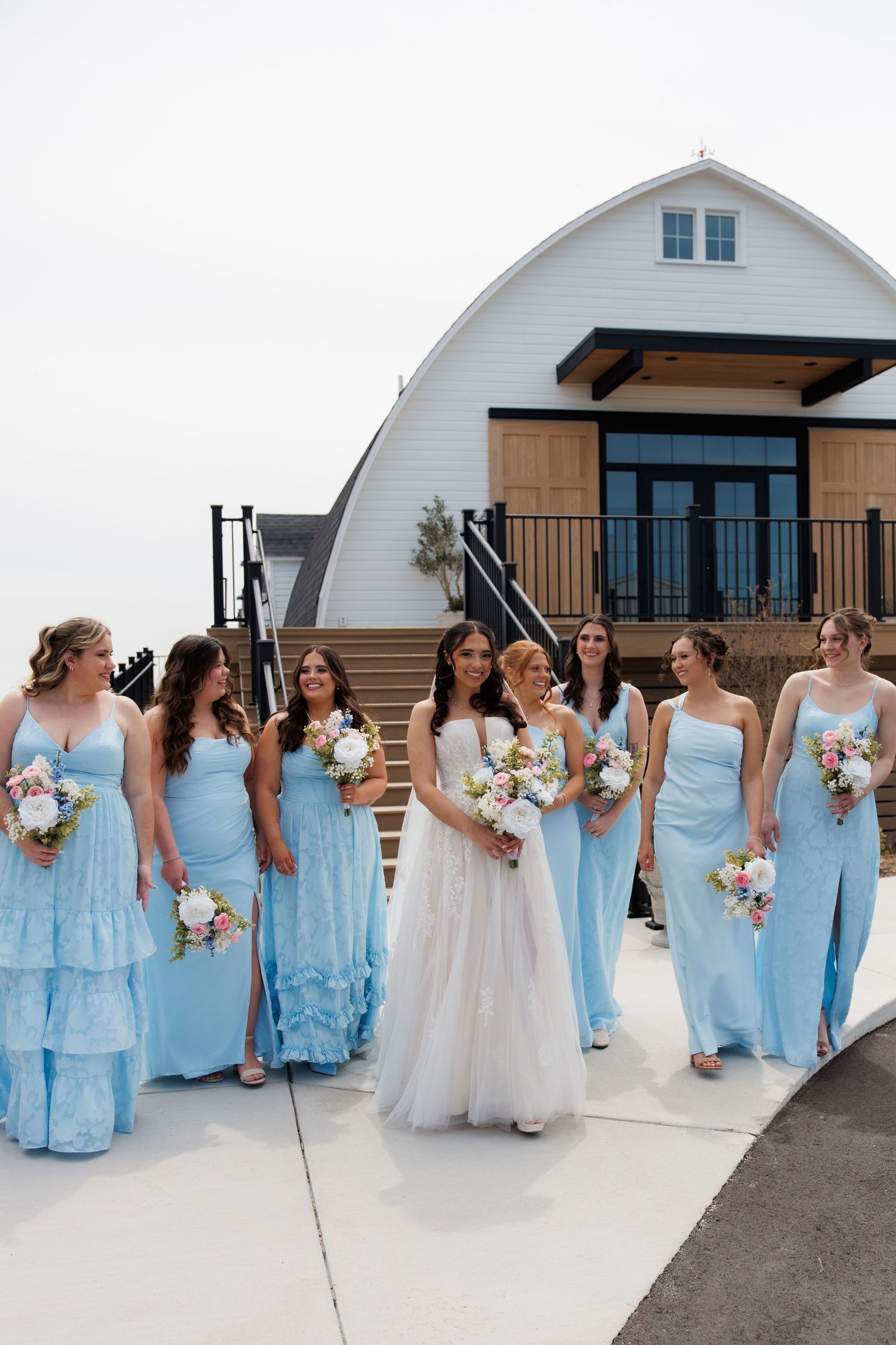 Bride and bridesmaids in light blue gowns walk together outside white barn wedding venue.