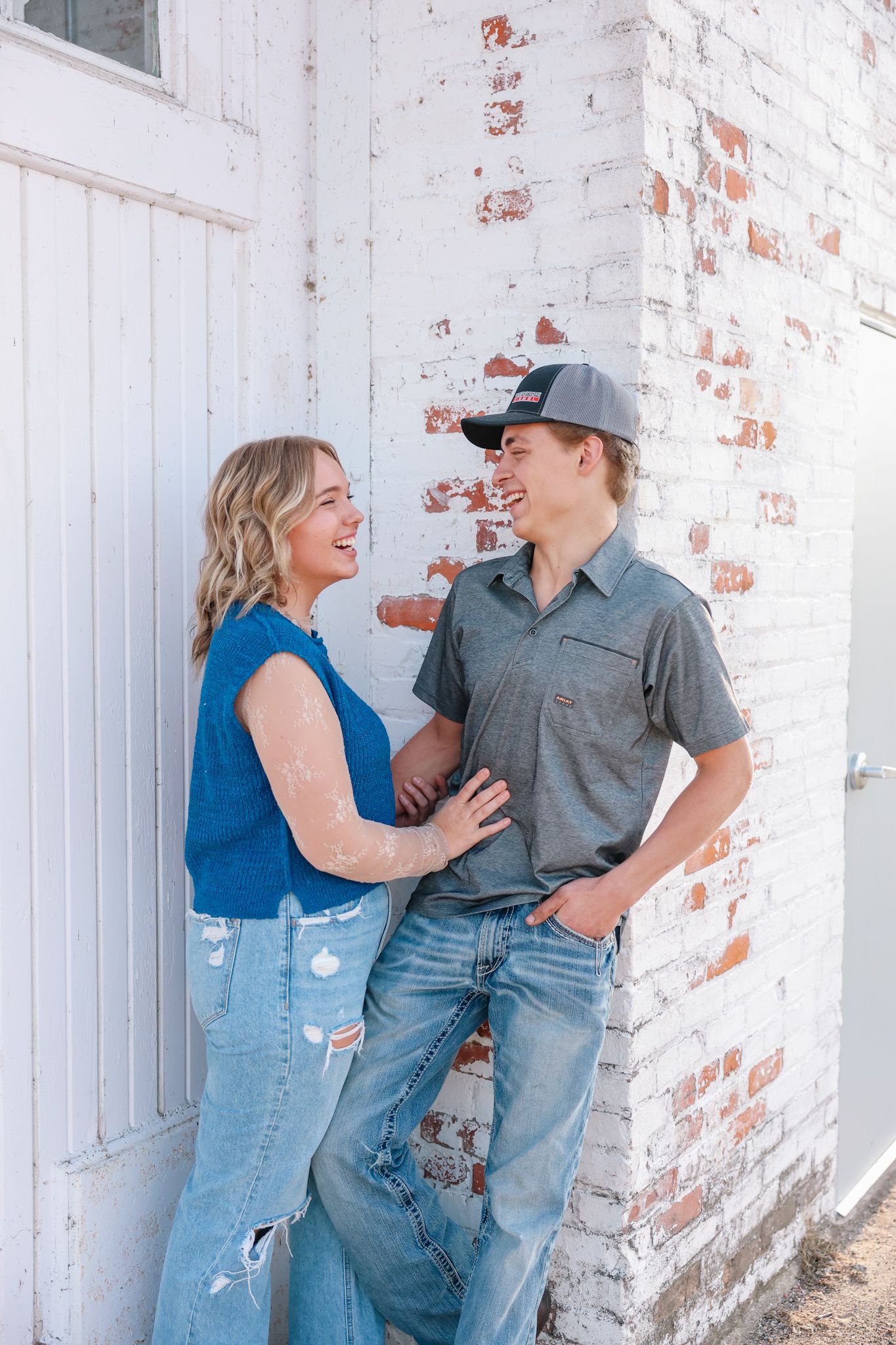 A laughing couple leans against a white brick wall, the woman in a blue top and the man in a gray shirt and cap.