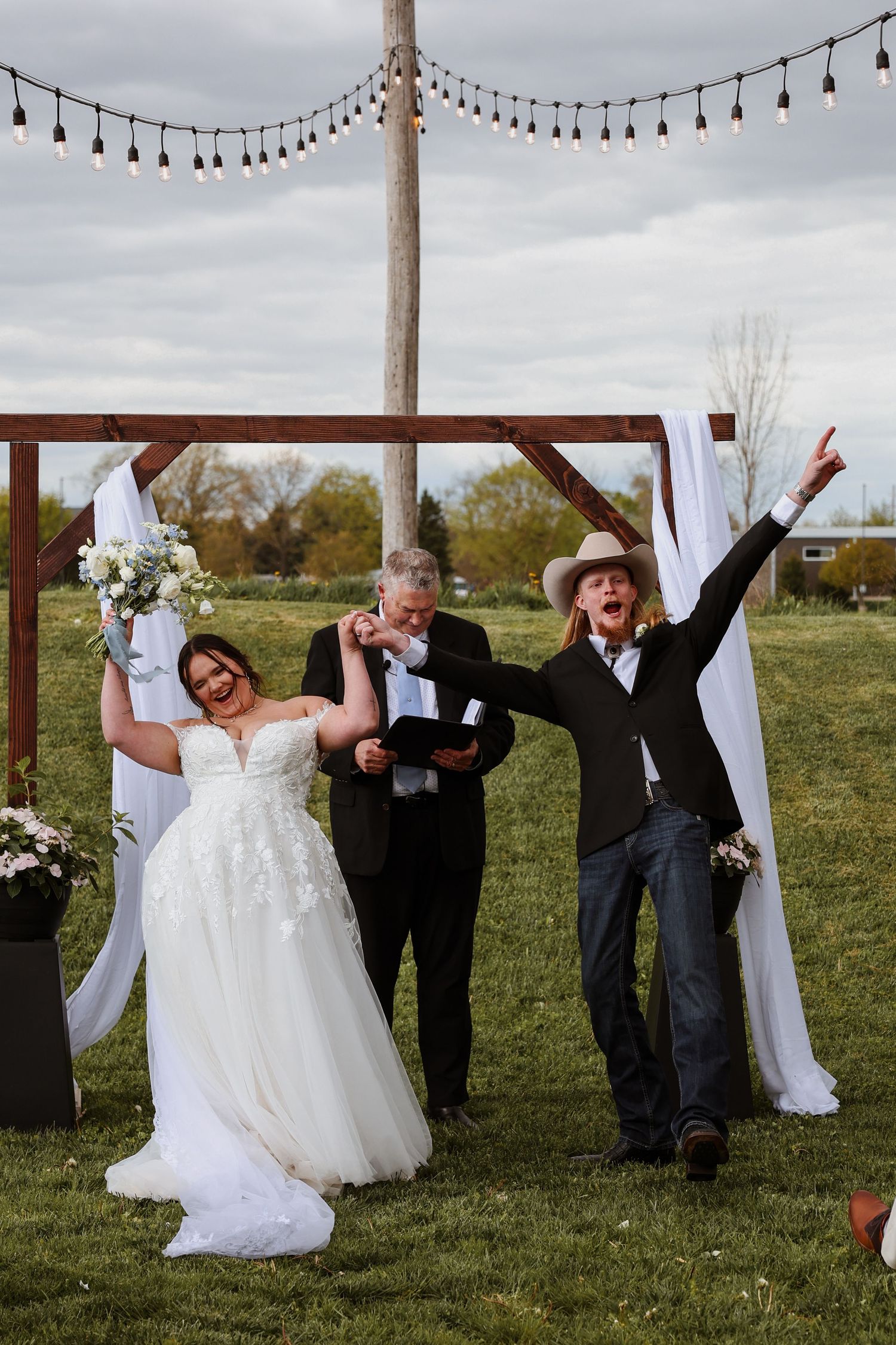 Joyful bride and groom celebrate after outdoor wedding ceremony under rustic wooden arch with string lights.