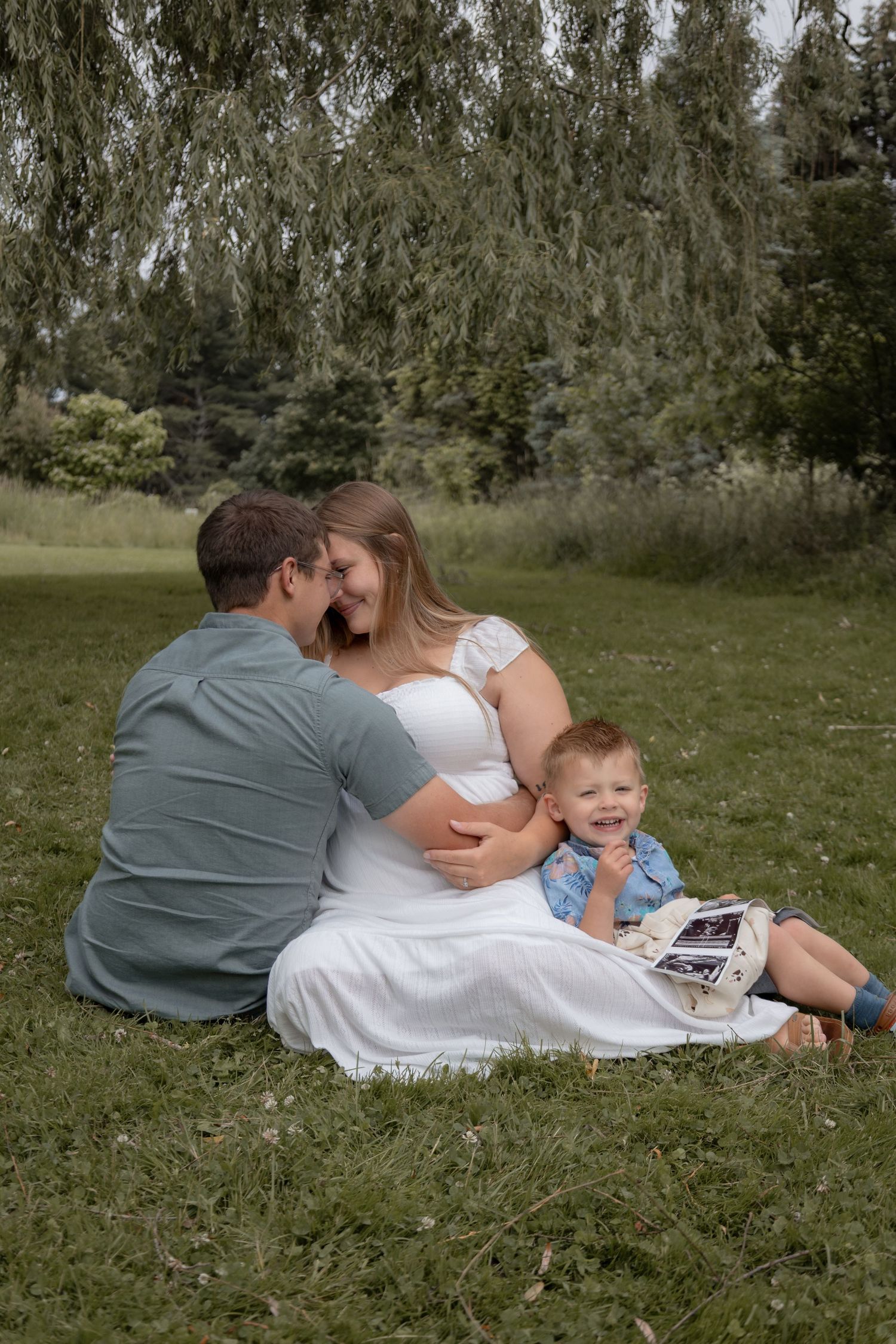 Pregnant couple sitting on grass with smiling toddler holding ultrasound photos under a weeping willow tree.