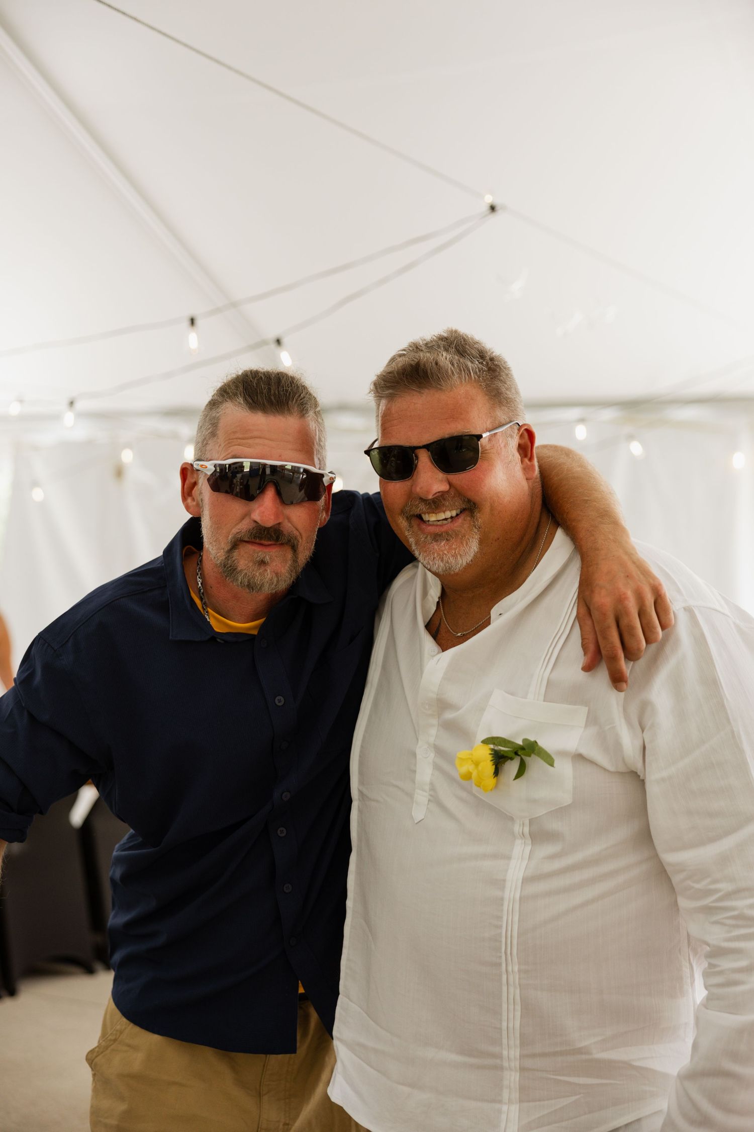 Two smiling men wearing sunglasses pose together under white tent at outdoor wedding reception.