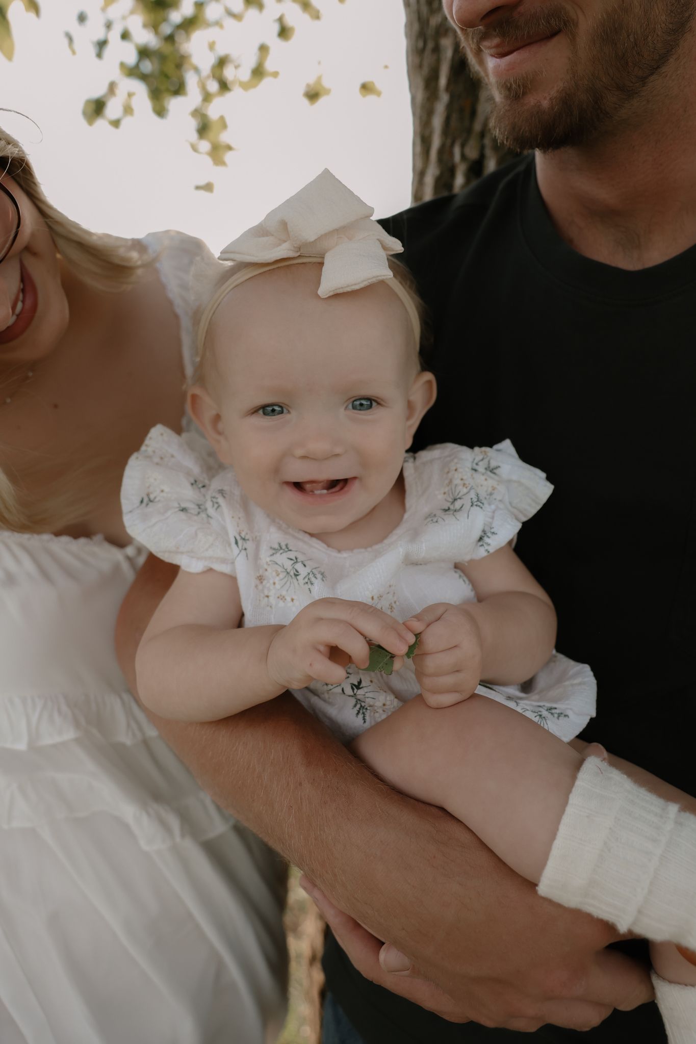 Adorable smiling baby girl in white floral romper with bow headband held by parents outdoors.