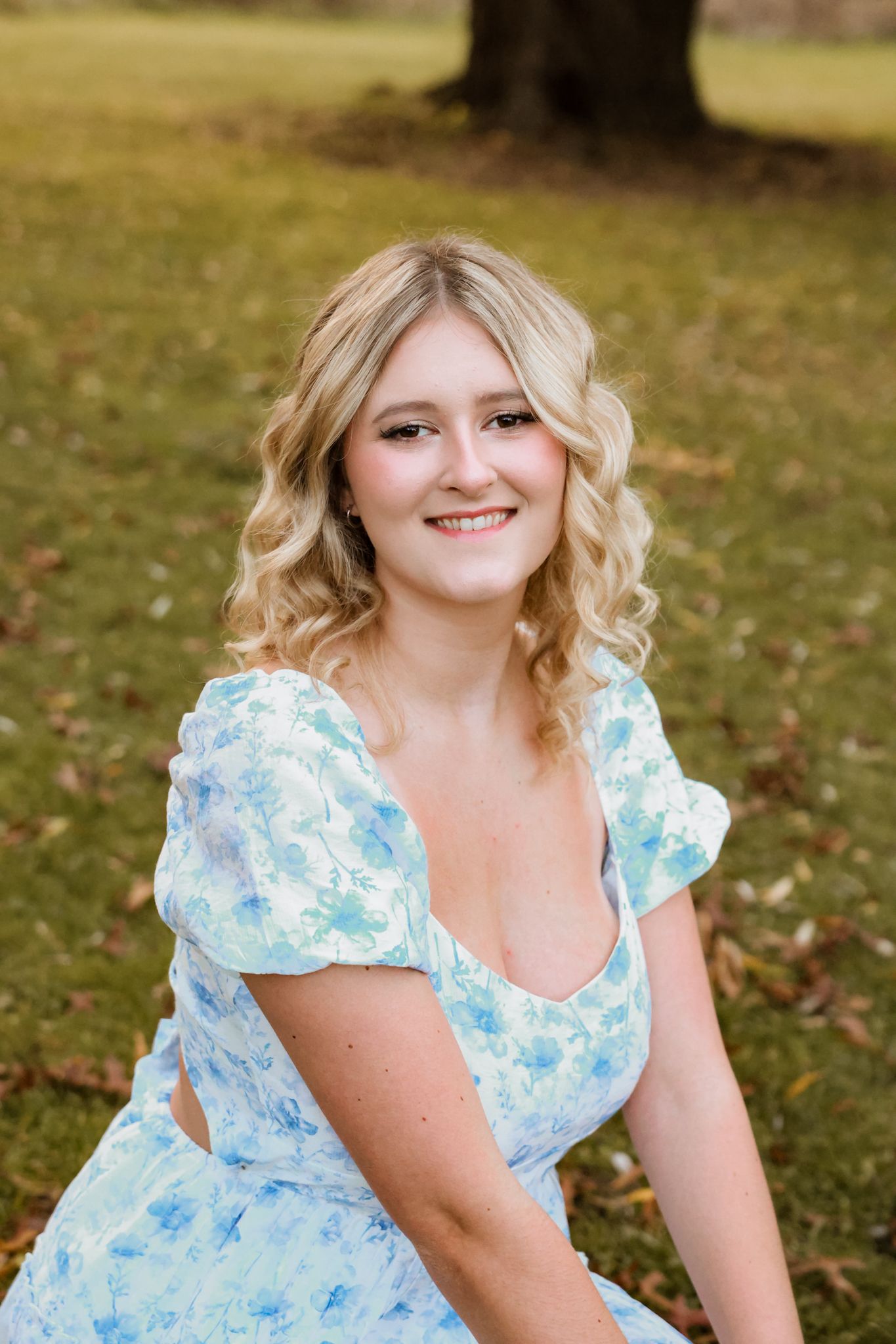 Smiling blonde woman in blue floral dress sitting outdoors in autumn park setting.