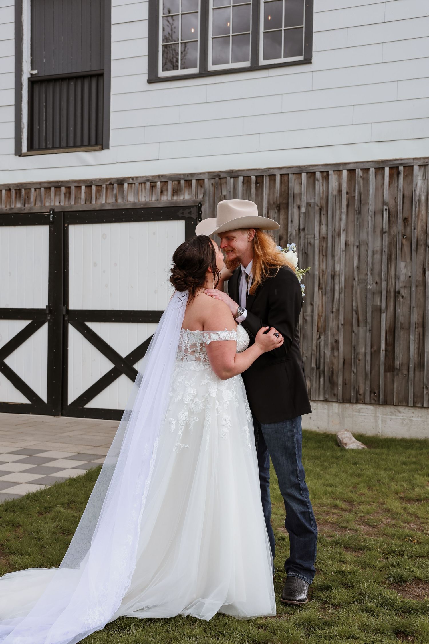 Bride and groom sharing a kiss outdoors, groom wearing cowboy hat and black blazer beside rustic barn.
