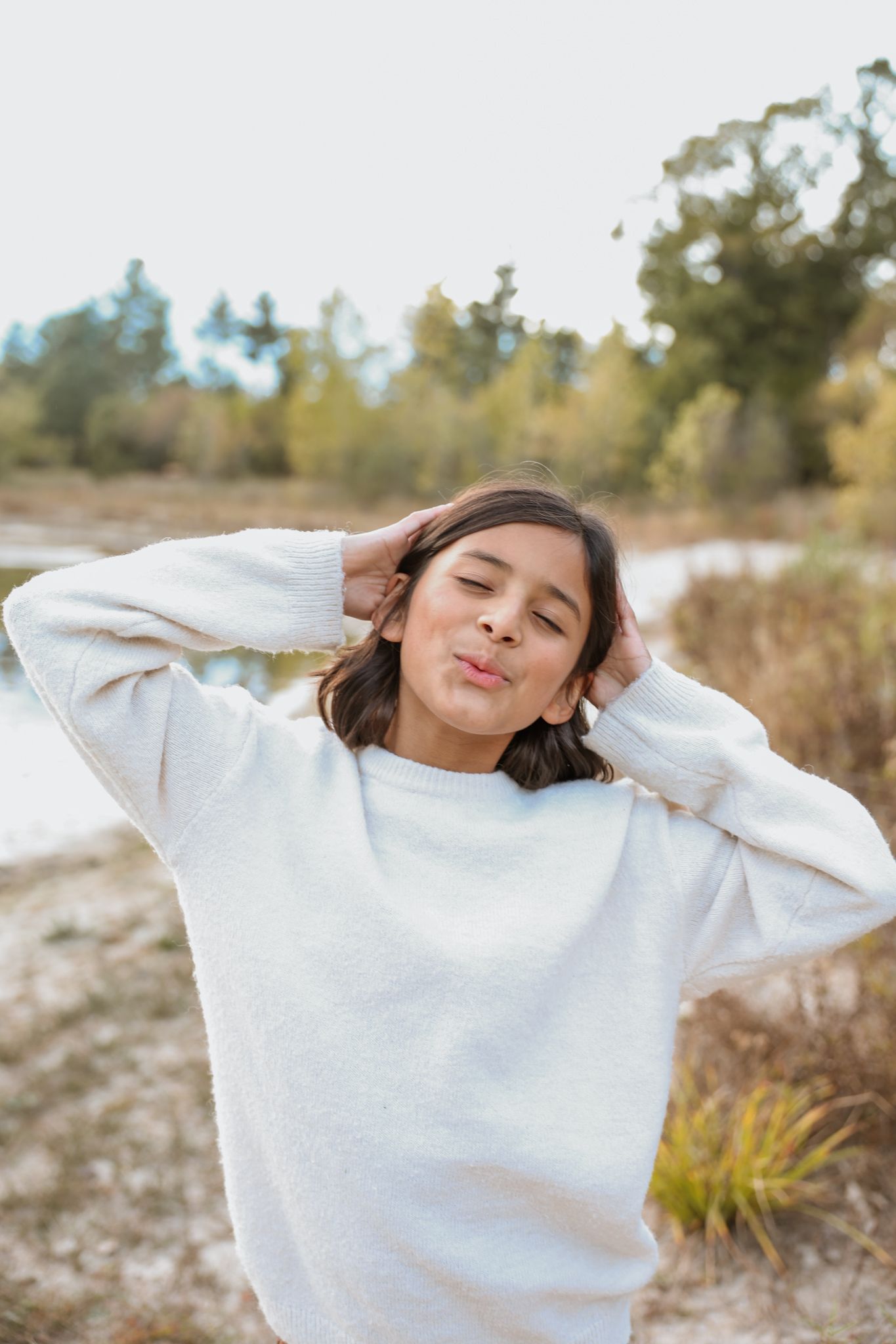 Young girl in white sweater makes a kissing face with hands behind her head outdoors.