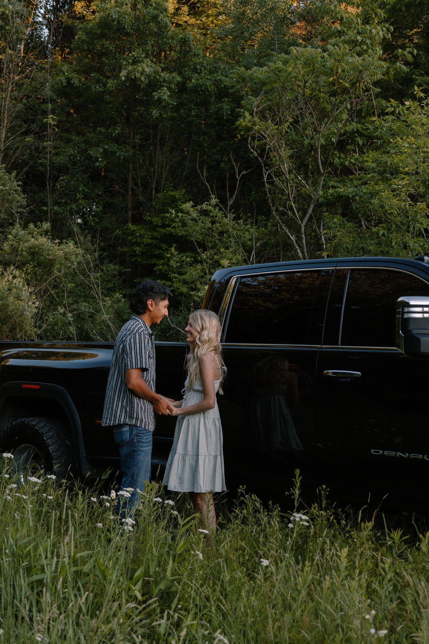 Couple holding hands near black GMC Denali truck surrounded by green forest.