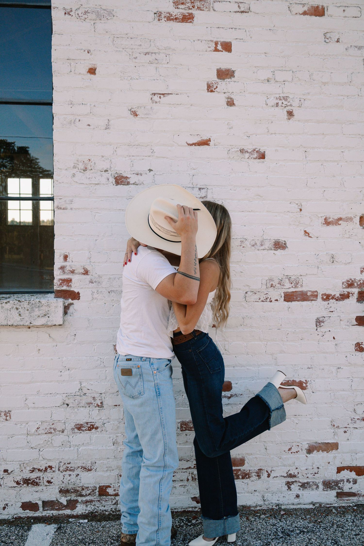 Couple kissing against white brick wall, woman in wide-brim hat and jeans, man in white tee and light jeans.