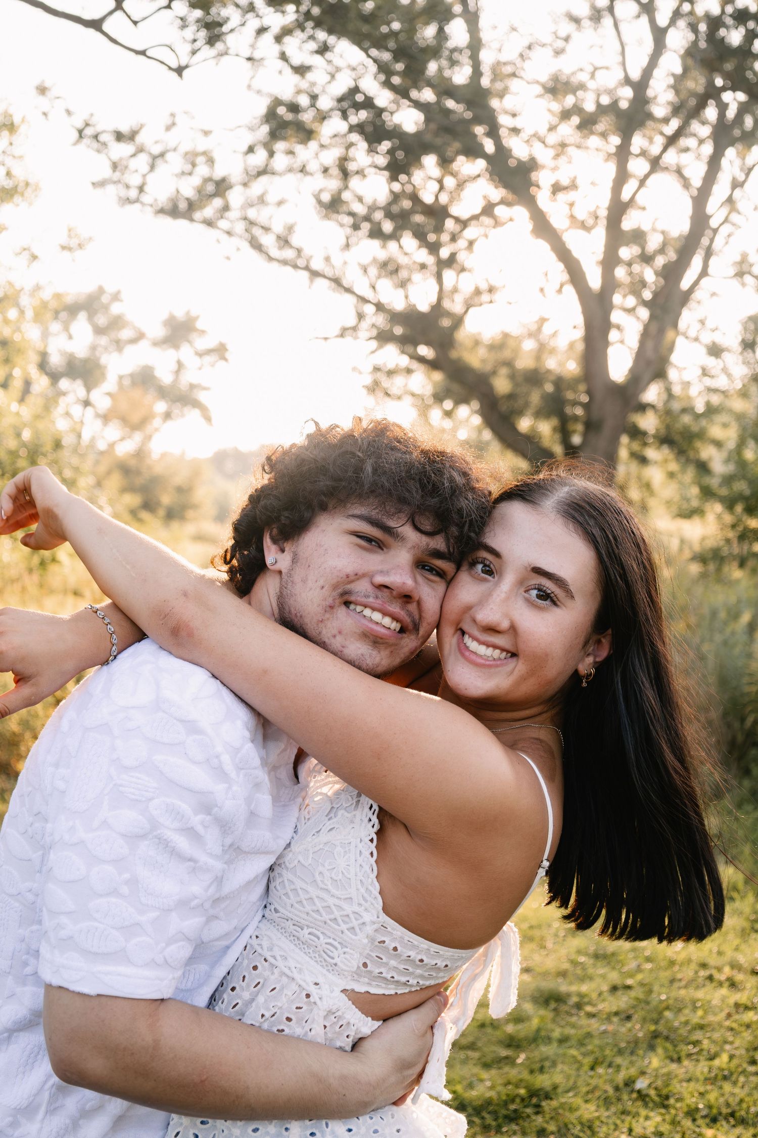 A smiling couple embraces outdoors in golden sunlight, woman in white dress hugging man from behind near green trees.
