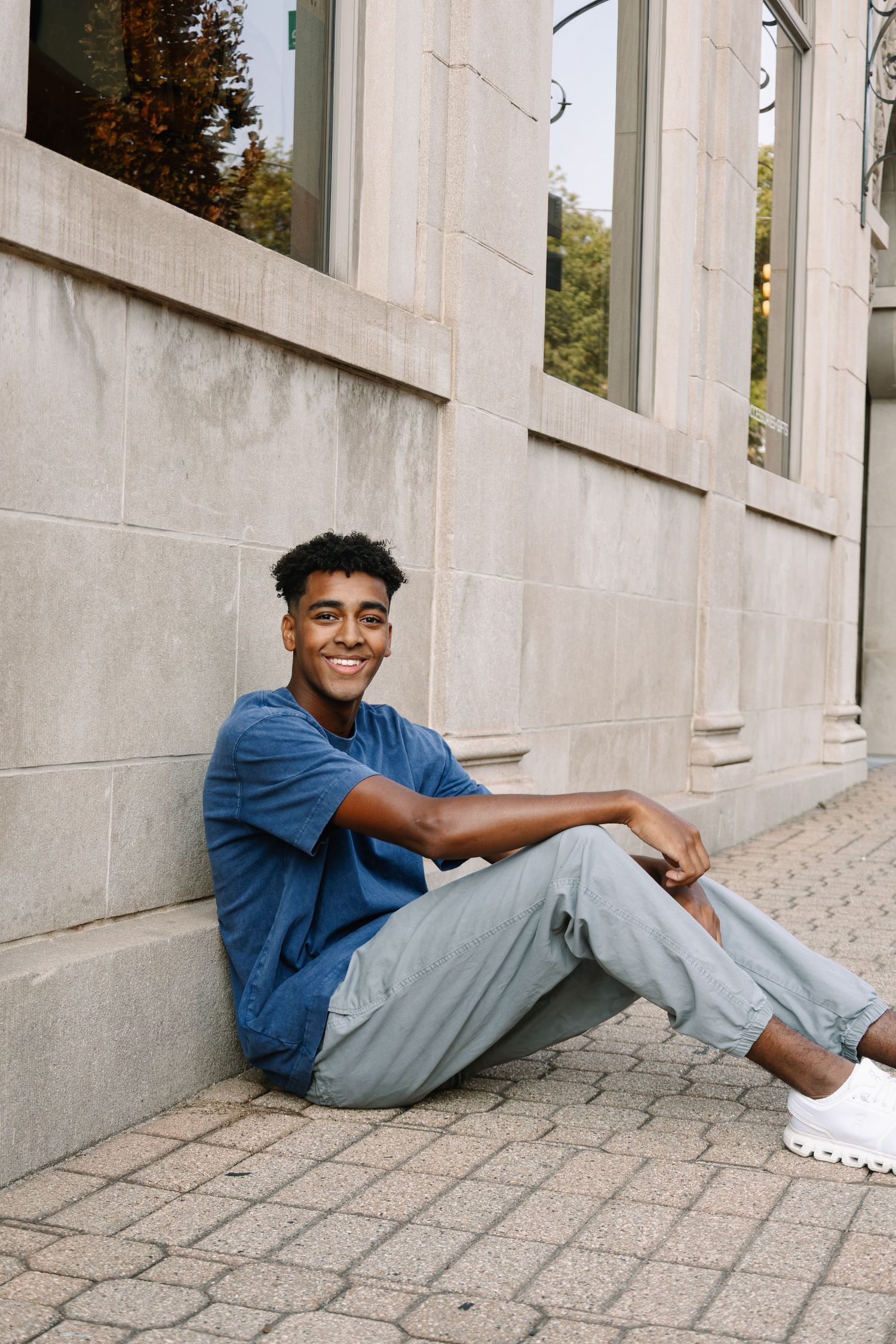 Young man in blue t-shirt and gray pants sitting against a stone building wall, smiling outdoors.