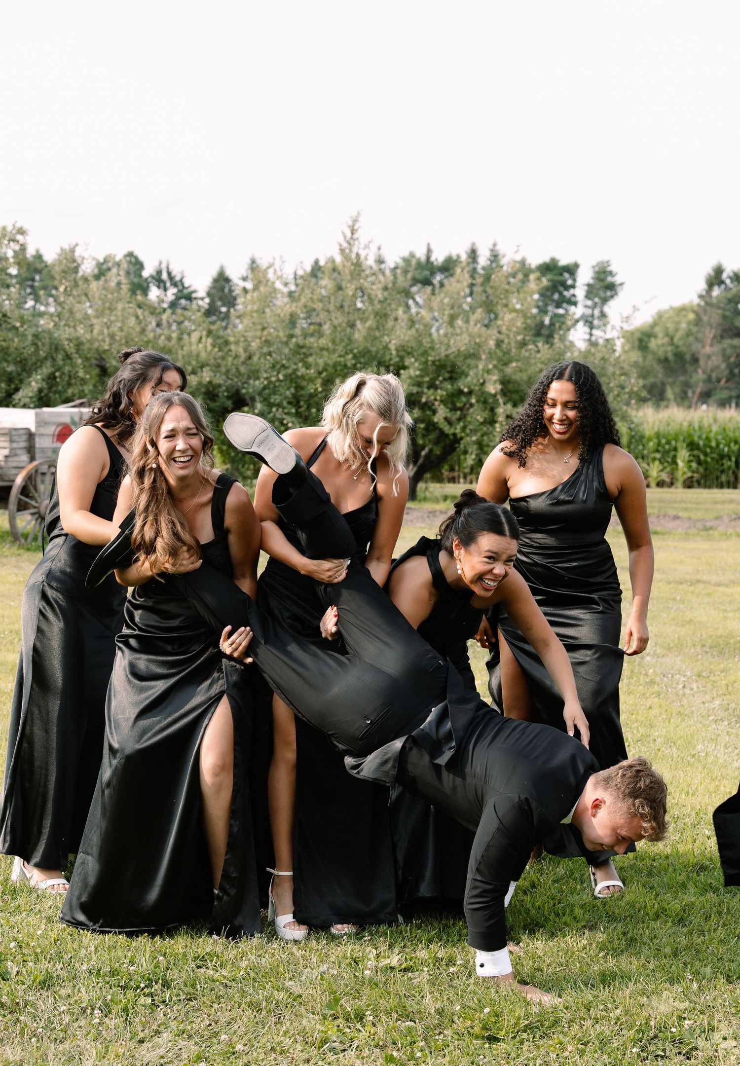 Bridesmaids in black gowns laughing outdoors as groomsman does push-ups on grass at wedding reception.
