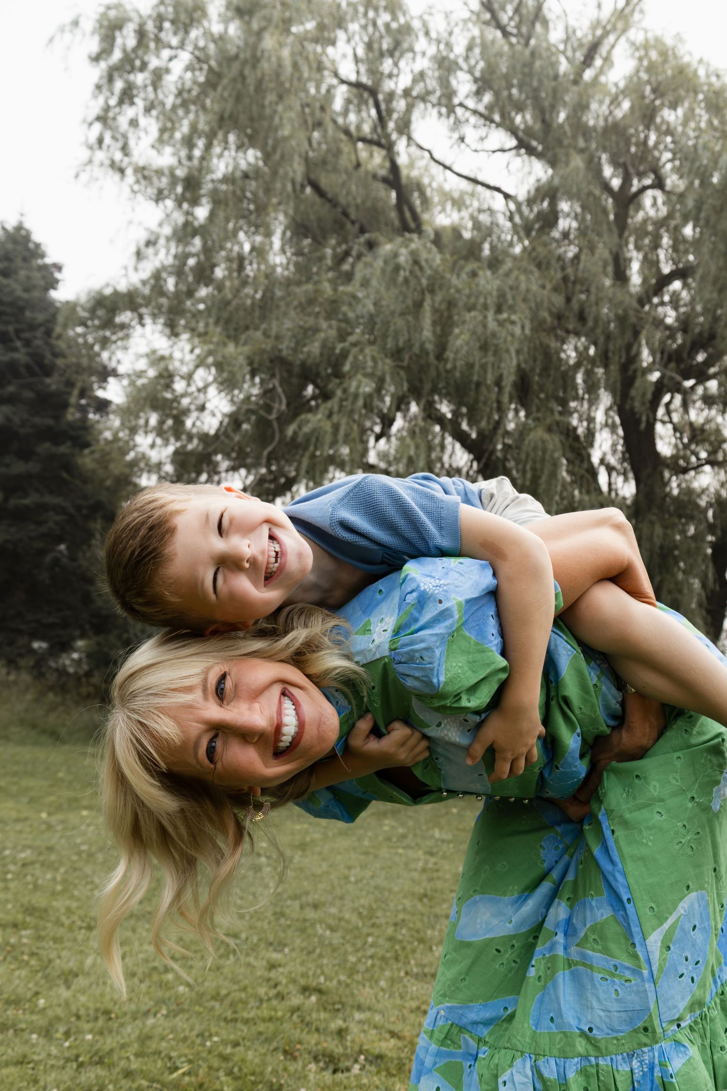 A smiling woman giving a happy child a piggyback ride outdoors in a green park with trees in the background.