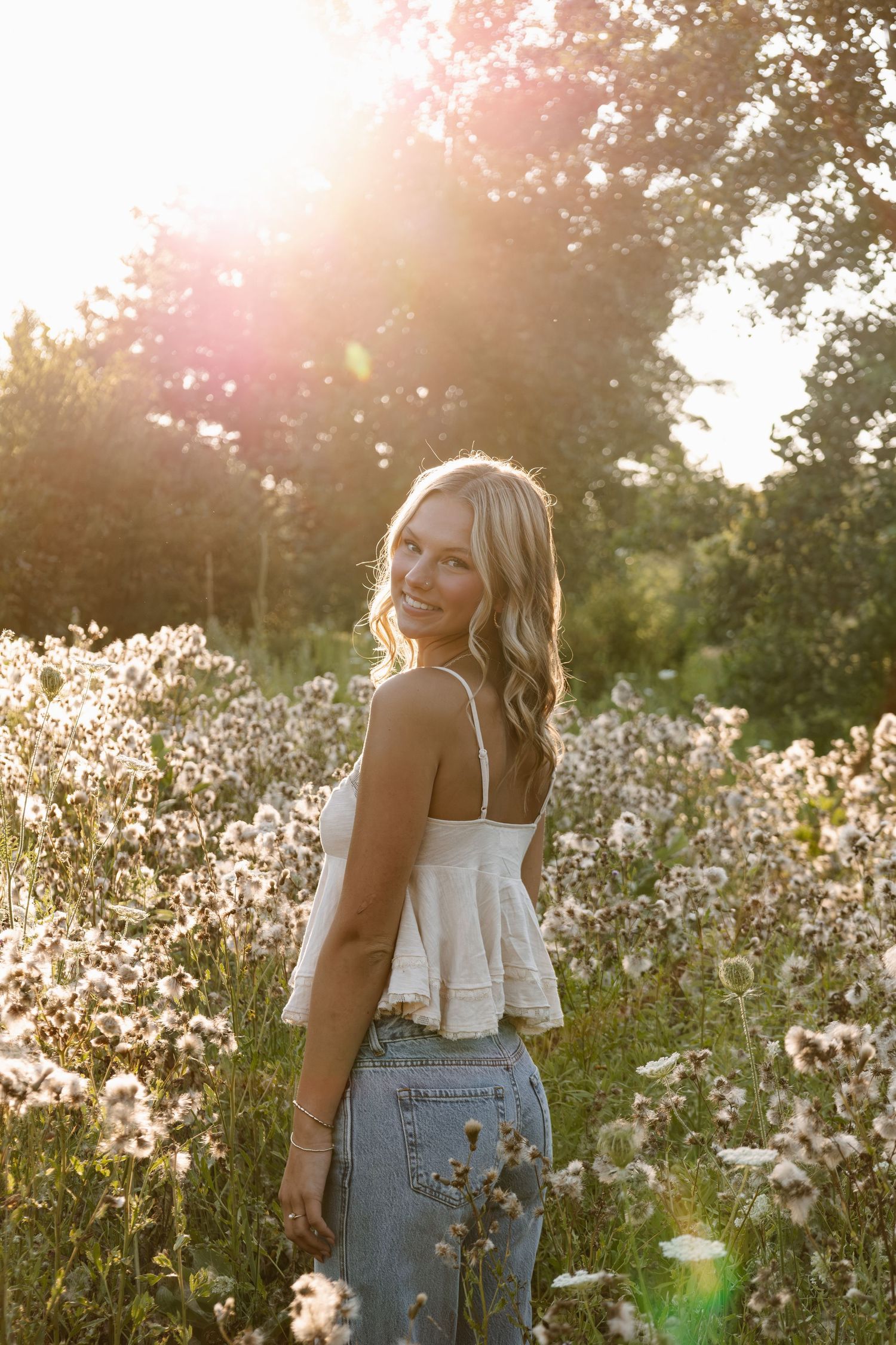 Smiling blonde woman in white crop top and jeans standing in a wildflower field with golden sunset light.