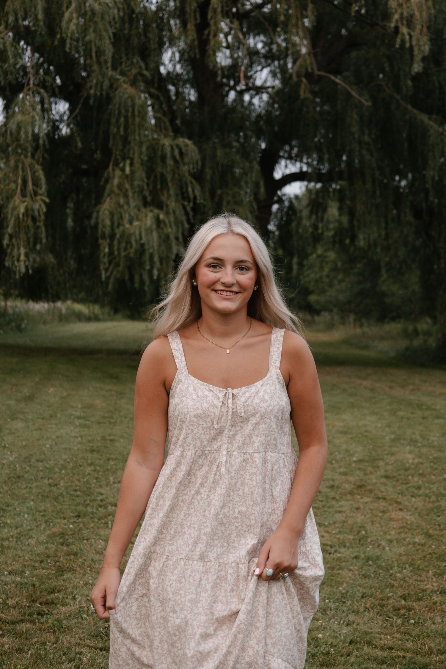 Smiling blonde woman in floral sundress standing outdoors near a weeping willow tree on a green lawn.