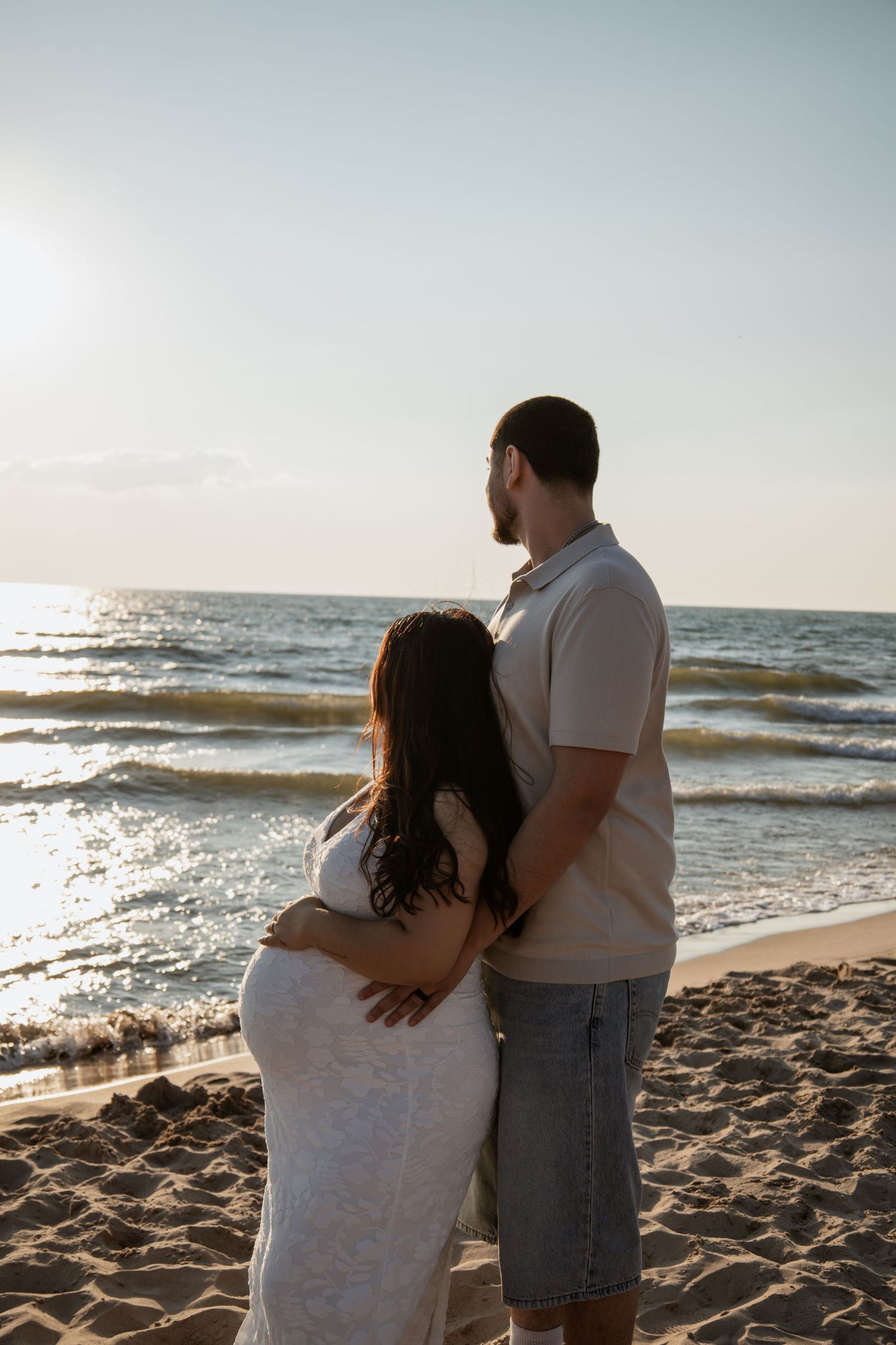 Pregnant couple embracing on a sandy beach at sunset, gazing at ocean waves in a romantic maternity photo.
