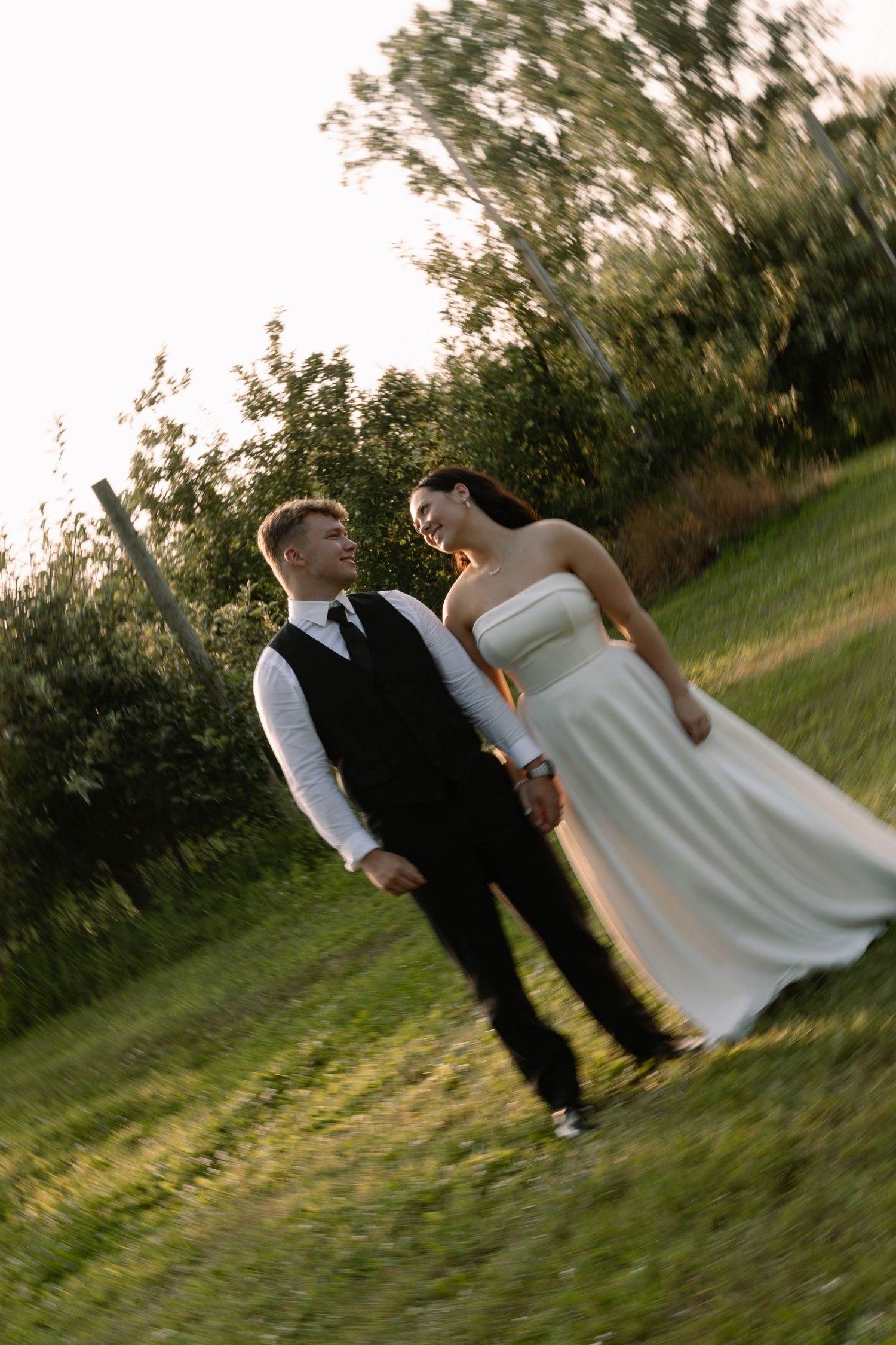 Bride and groom walk hand-in-hand through lush green orchard during golden hour wedding portraits.