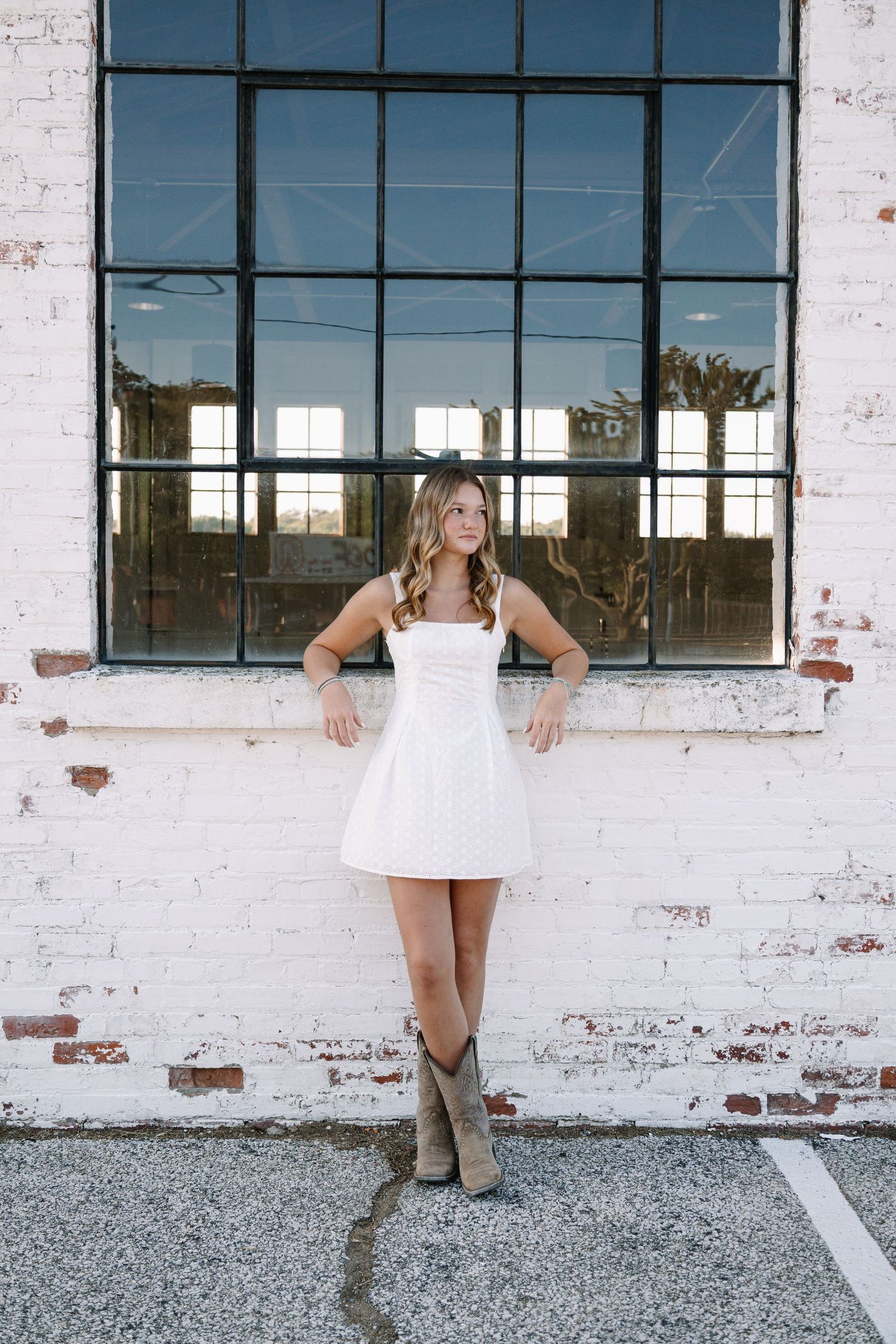 Young woman in white dress and cowboy boots leaning against white brick wall outdoors.