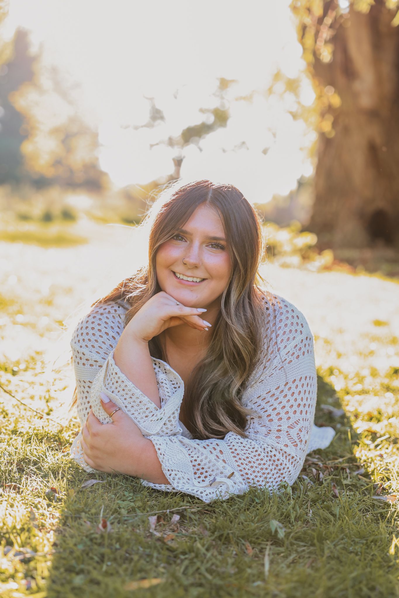 Smiling young woman in crochet cardigan lying on grass in golden sunlight near a tree.