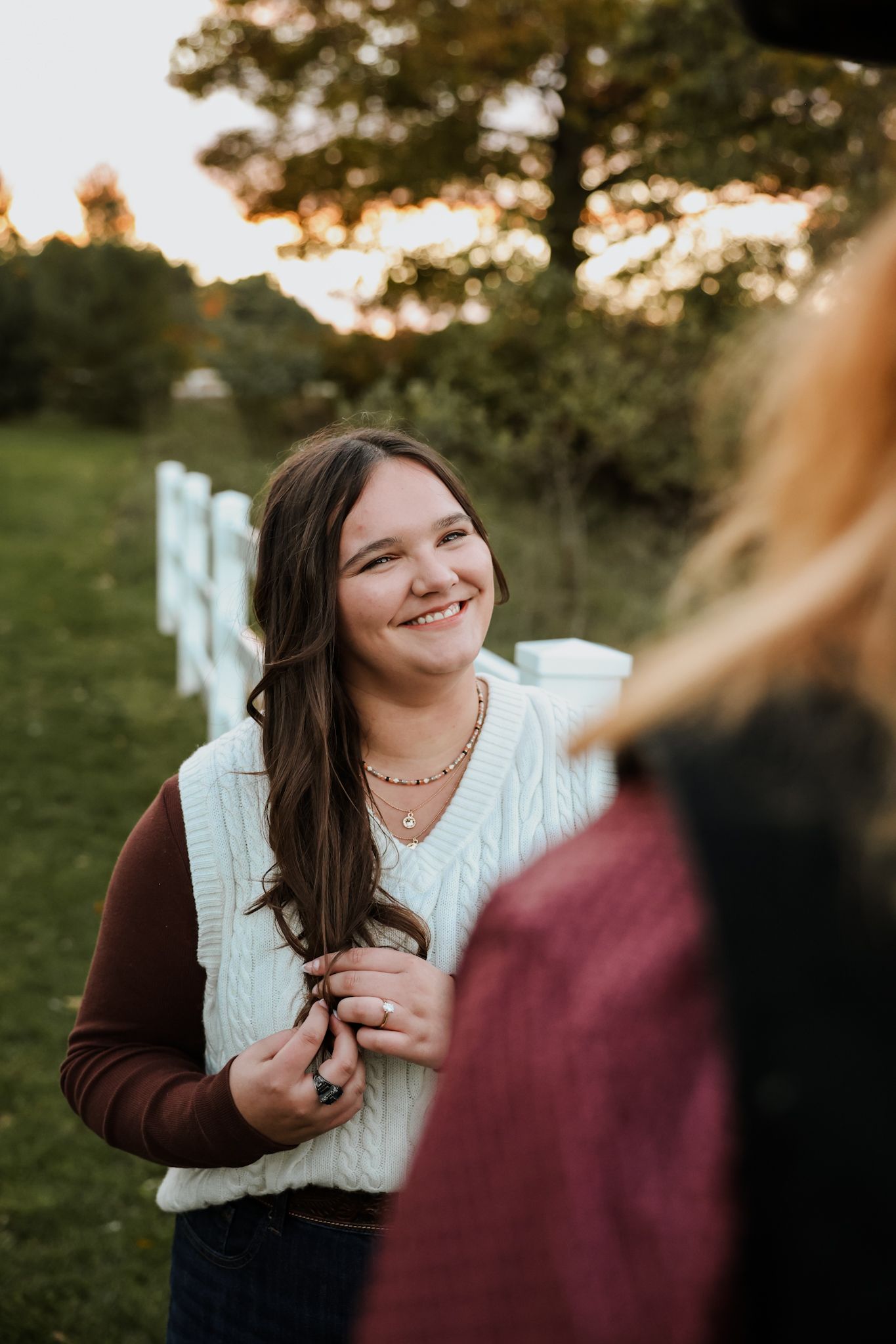 Young woman smiling warmly outdoors near white fence at golden hour in autumn.