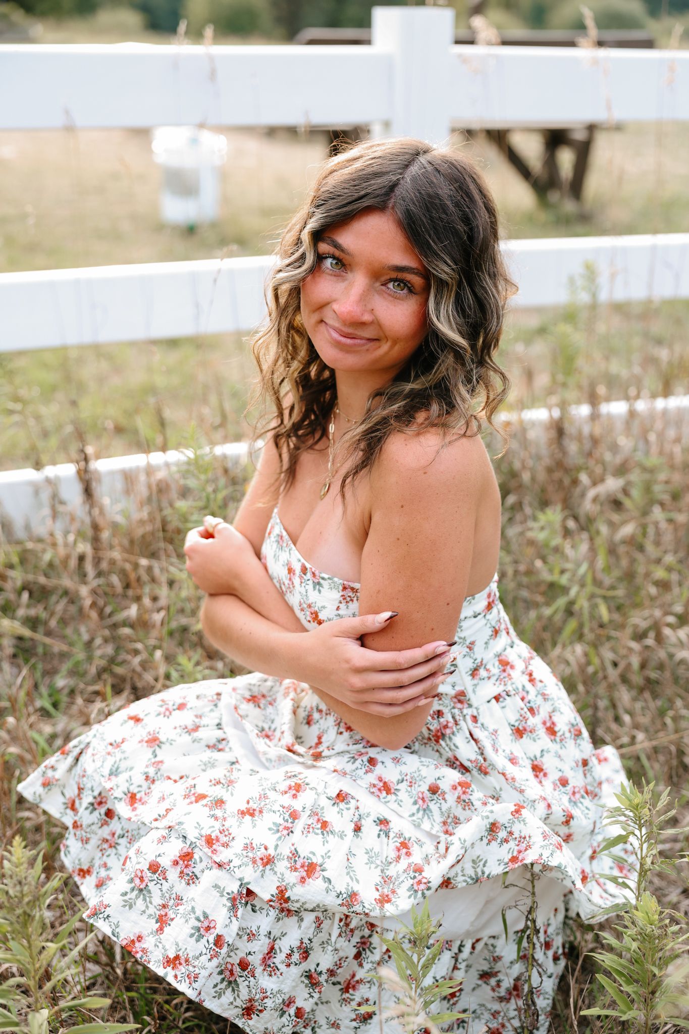 Young woman in floral dress sitting in field near white fence.
