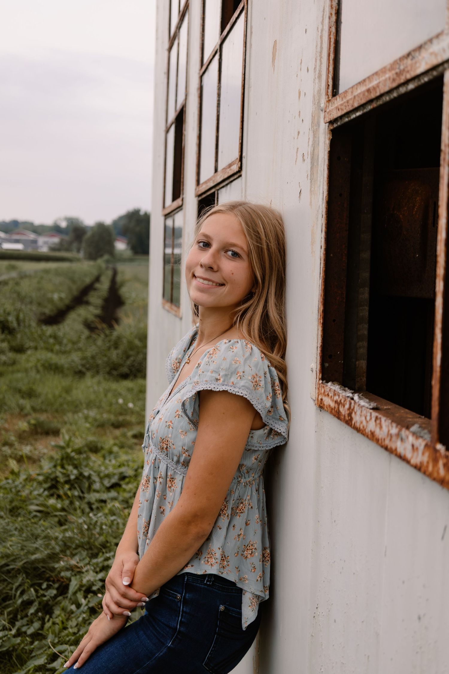 Young blonde woman in floral top and jeans leaning against rustic building with farm field backdrop.