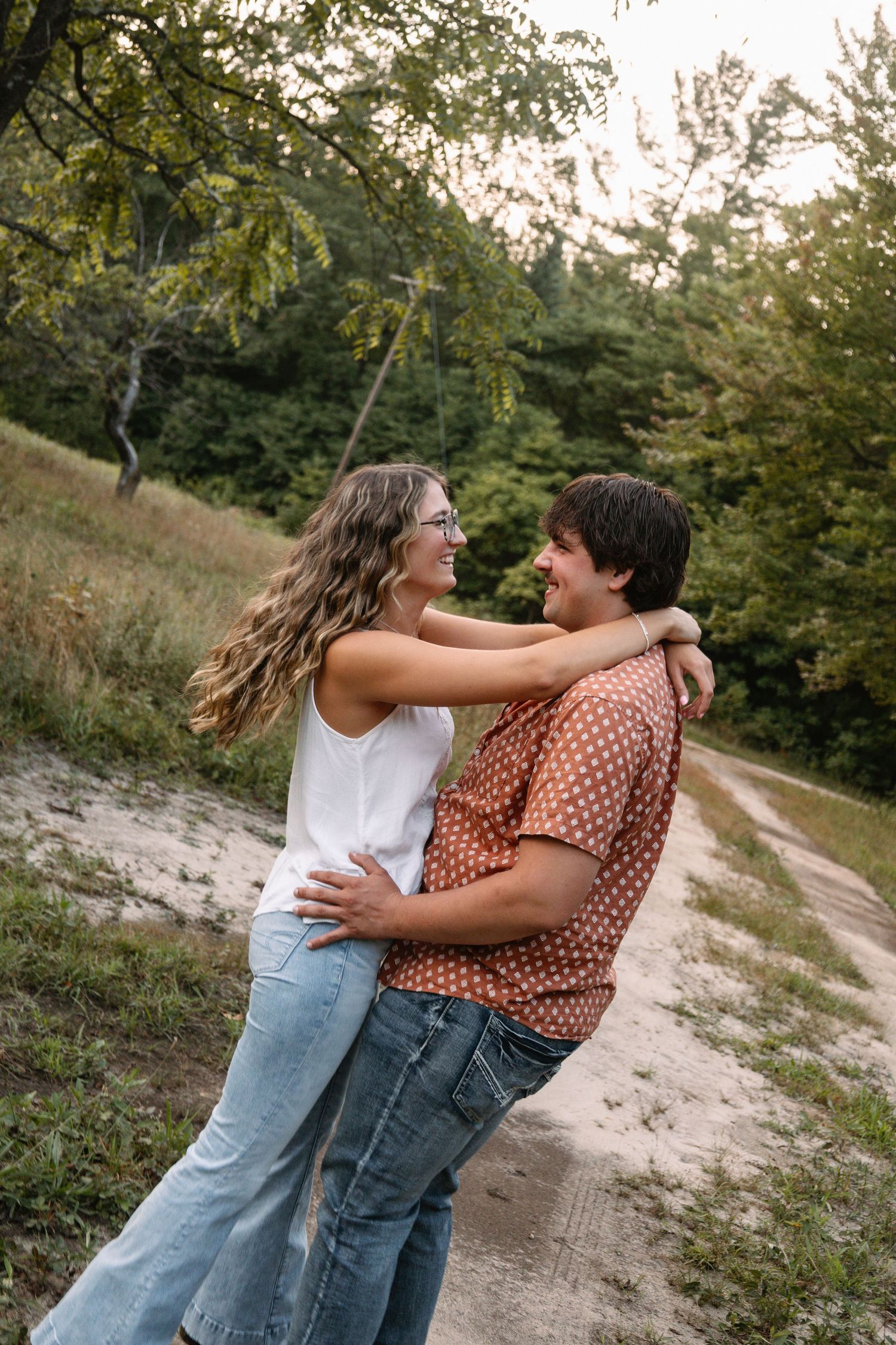 A happy couple embraces and smiles at each other on a sandy path surrounded by lush green trees.