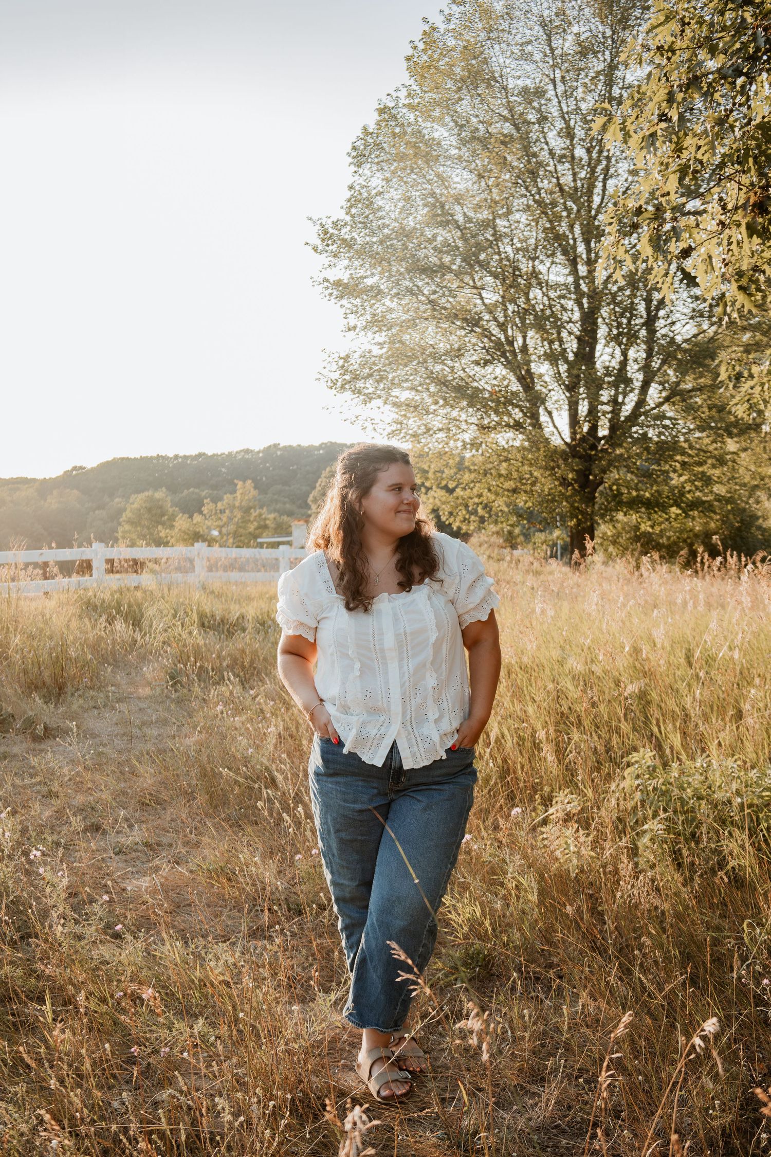 Woman in white blouse and jeans standing in golden field with trees and white fence in warm sunset light.
