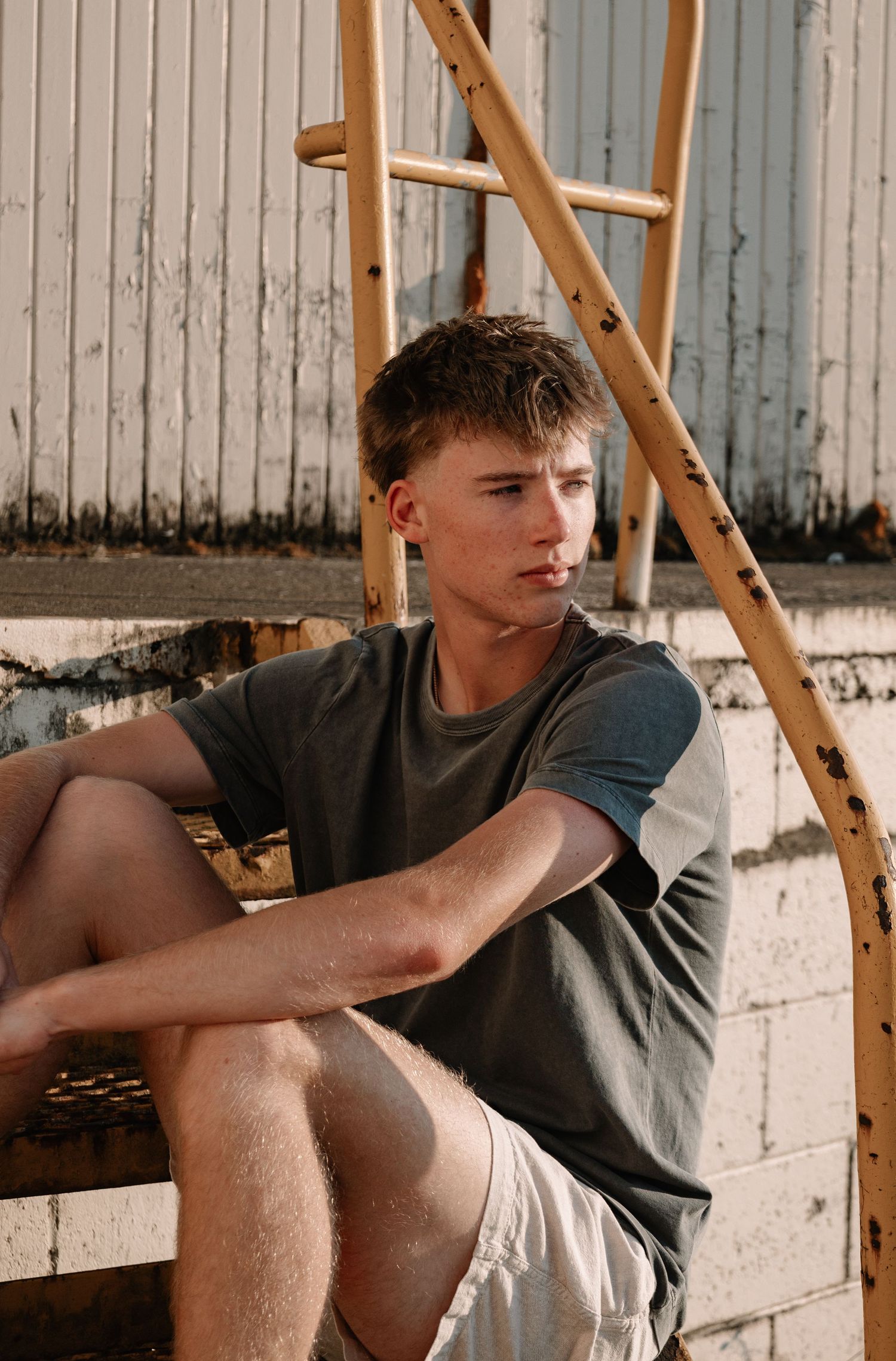 Young man in gray t-shirt and shorts sitting by a rustic ladder against a weathered white wall outdoors.