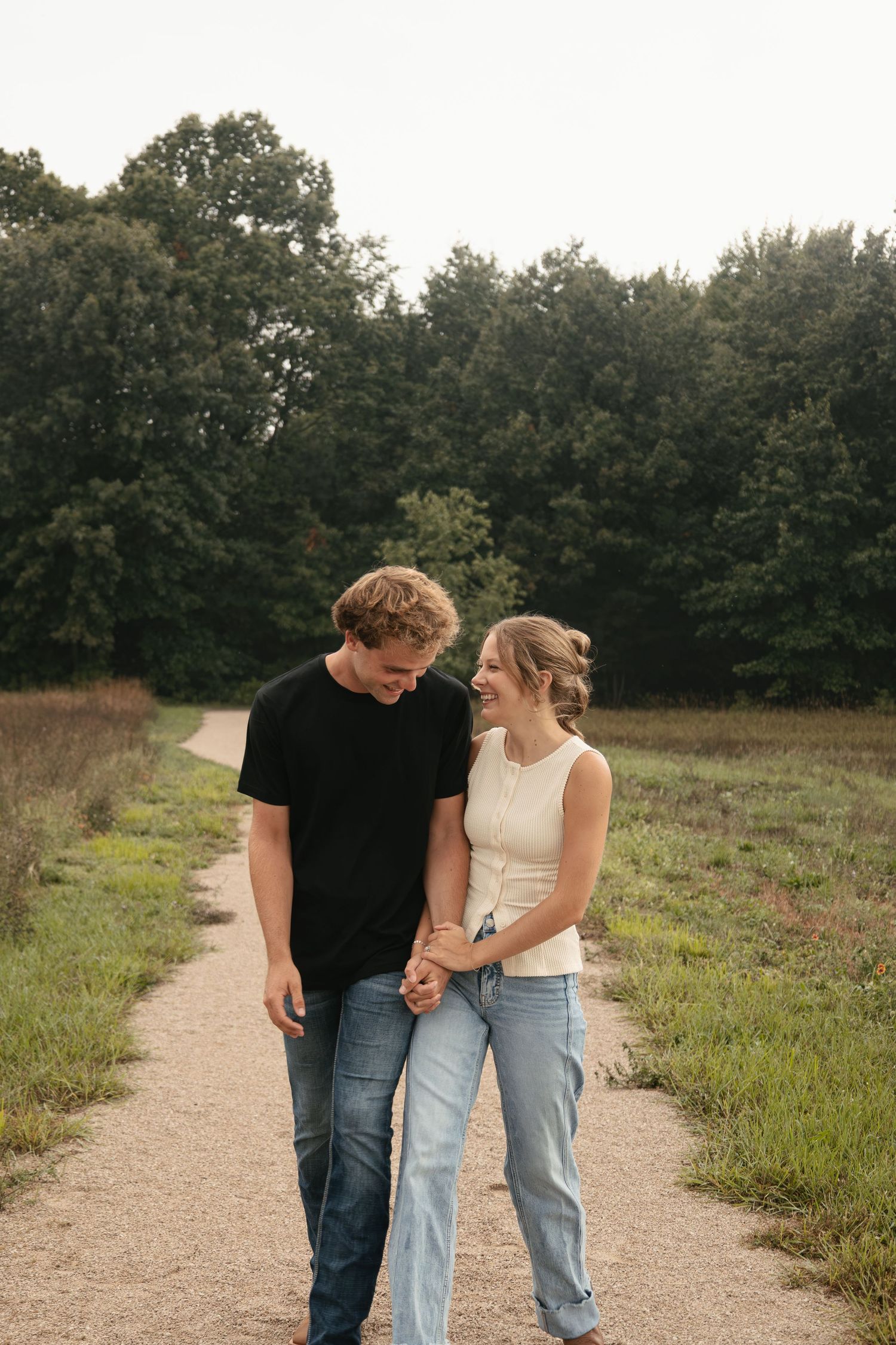A young couple holds hands and smiles at each other while walking on a path surrounded by green fields and trees.