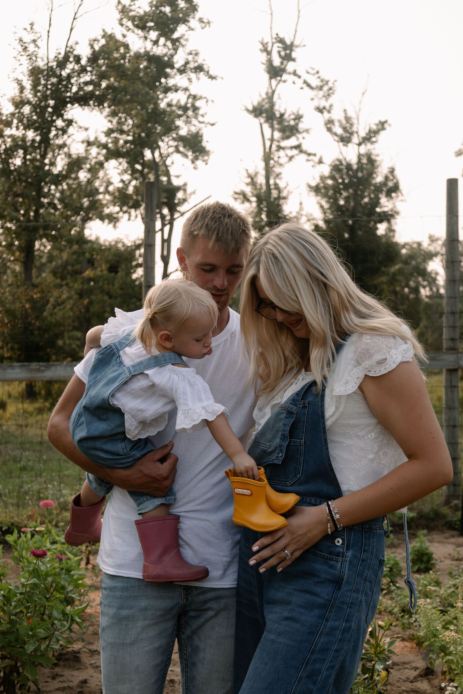 A young family with a toddler girl in denim overalls and rain boots enjoying an outdoor garden setting at golden hour.