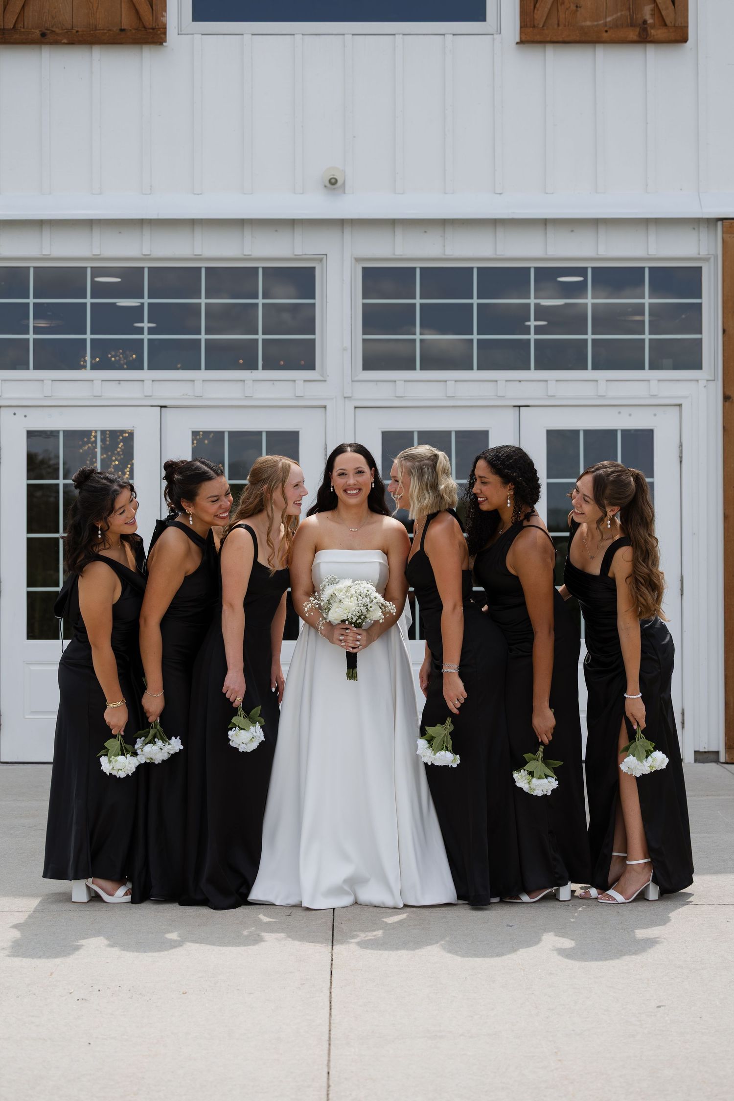 Bride in white gown holds bouquet surrounded by bridesmaids in black dresses outside a white barn venue.
