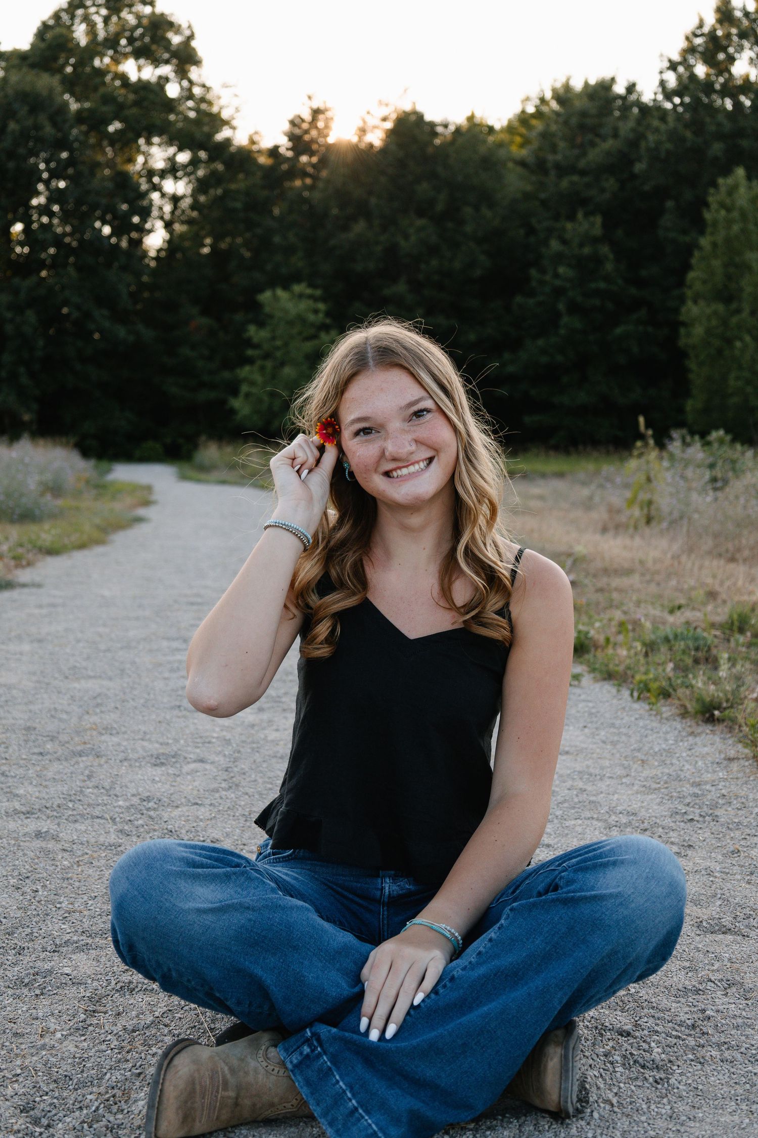Young woman sitting cross-legged on a gravel path, smiling, wearing a black tank top and jeans, with trees at sunset.
