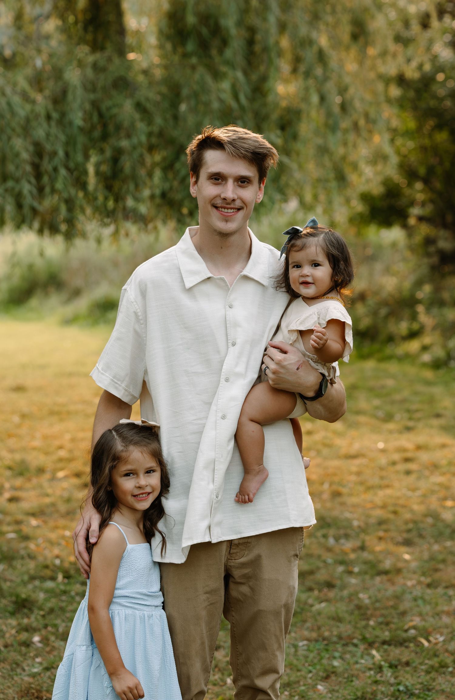 Young man smiling outdoors holding a toddler girl while a young girl in a blue dress stands beside him.