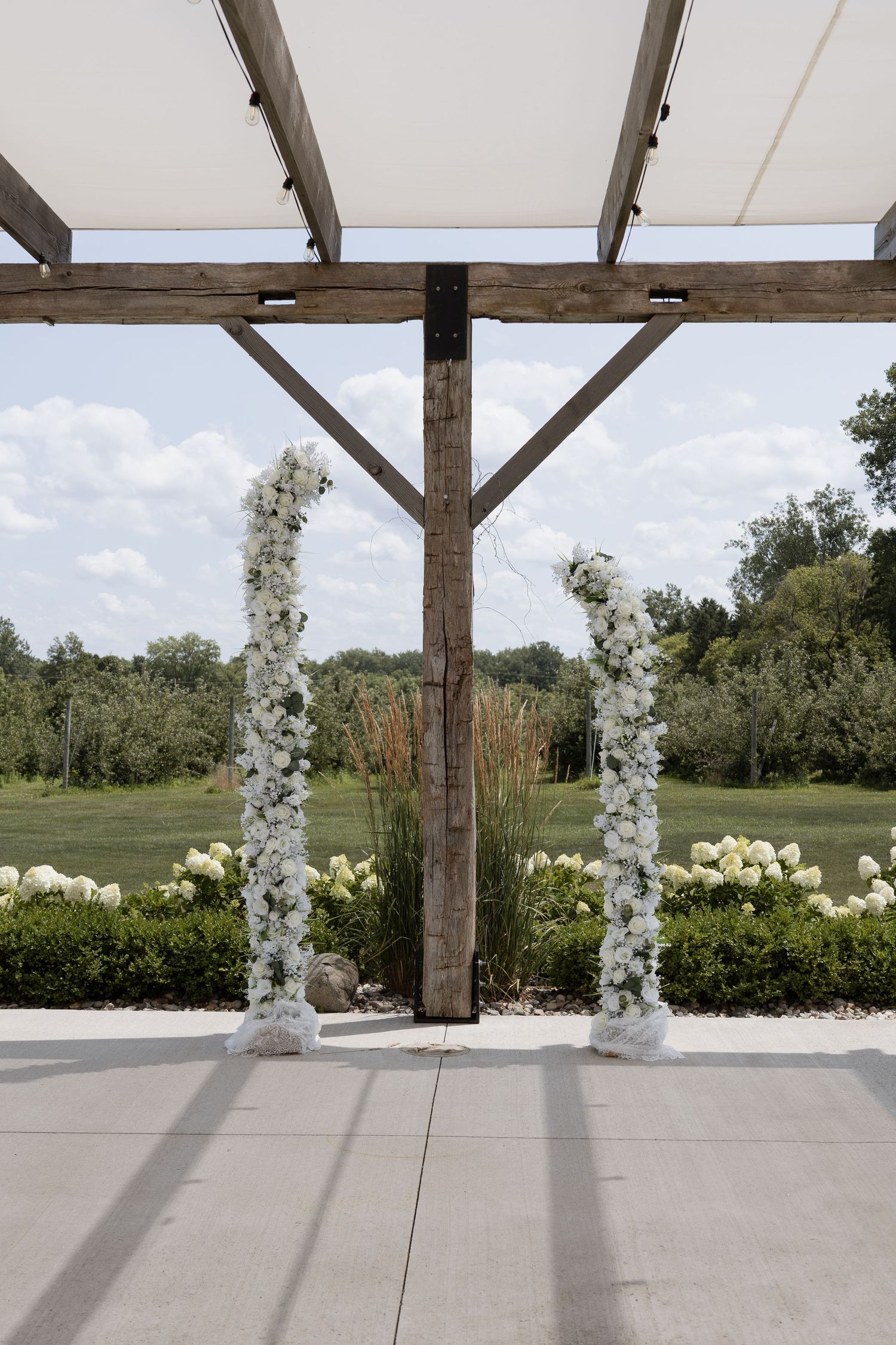 Elegant outdoor wedding ceremony arch with white floral columns framing a rustic wooden pergola overlooking a lush green landscape.
